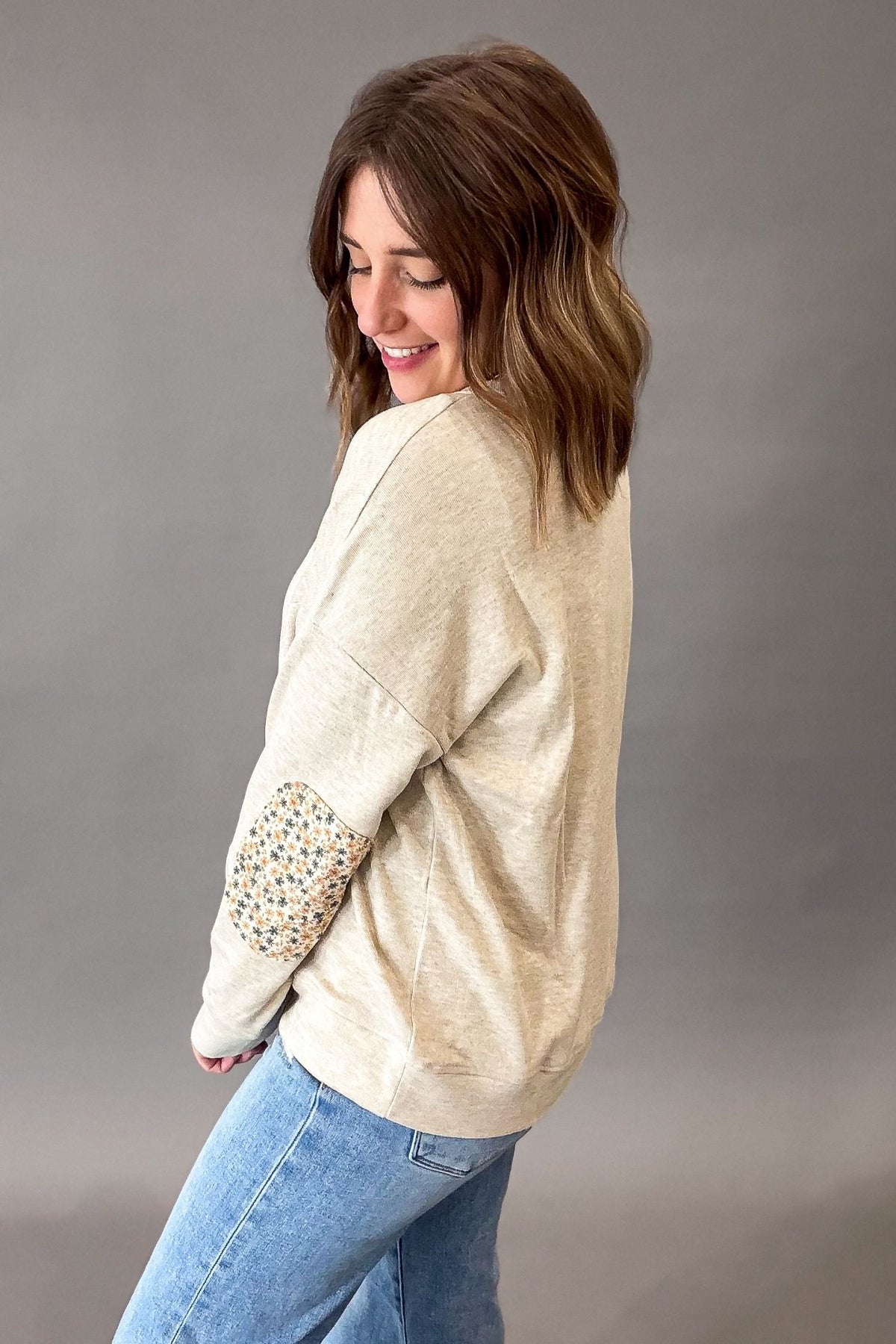 A woman wearing a oatmeal-colored knit sweatshirt with a floral patchwork elbow patch and blue jeans, photographed against a neutral gray studio background.