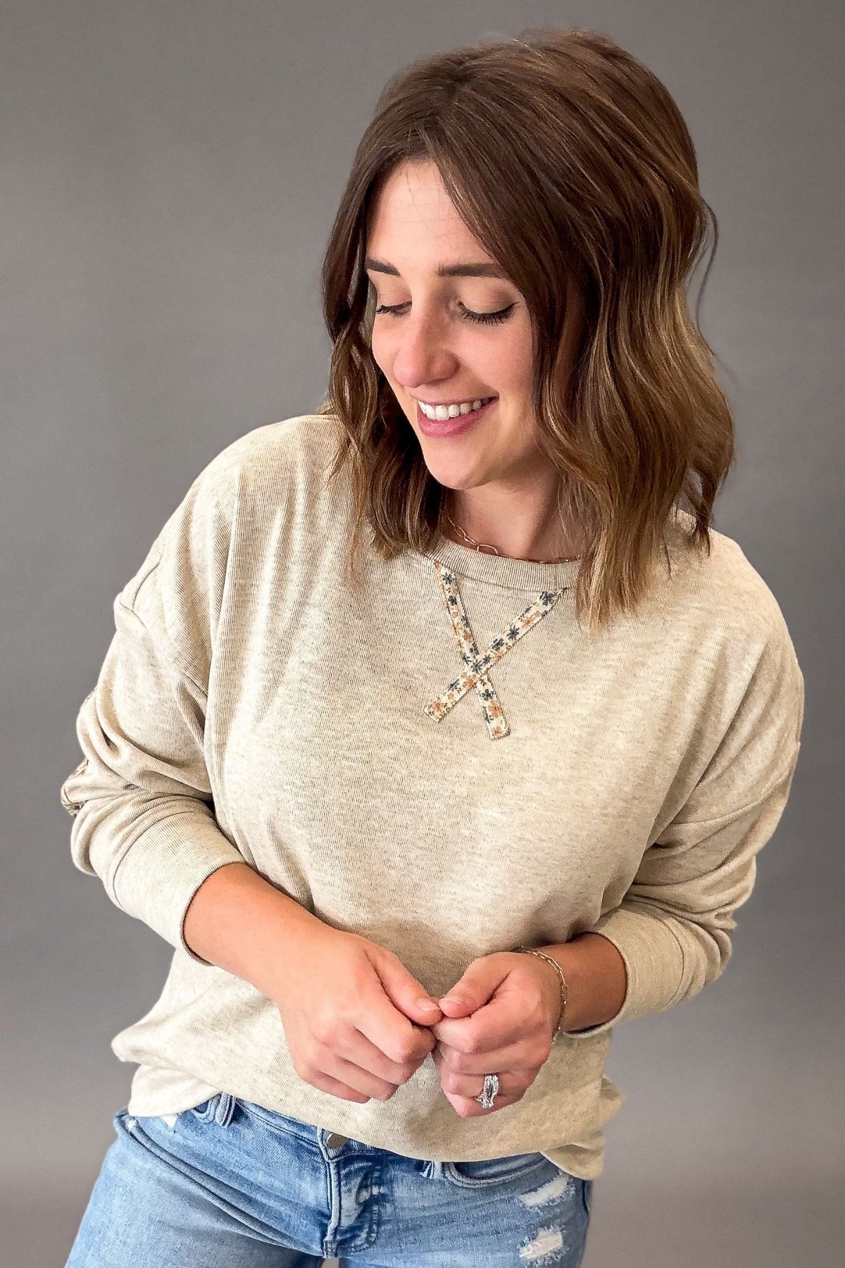 A woman with shoulder-length brown hair is shown from the waist up, wearing a light oatmeal long-sleeve knit top with a floral accent X-shaped neckline detail, paired with blue jeans, against a solid gray studio background.