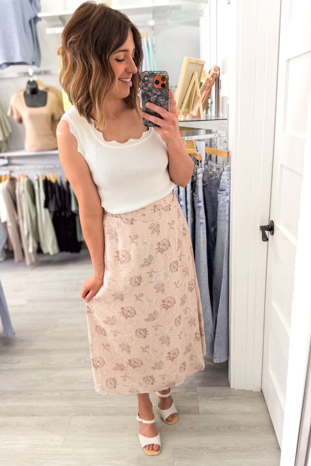Woman taking a mirror selfie wearing a white top and floral midi skirt in a clothing store.