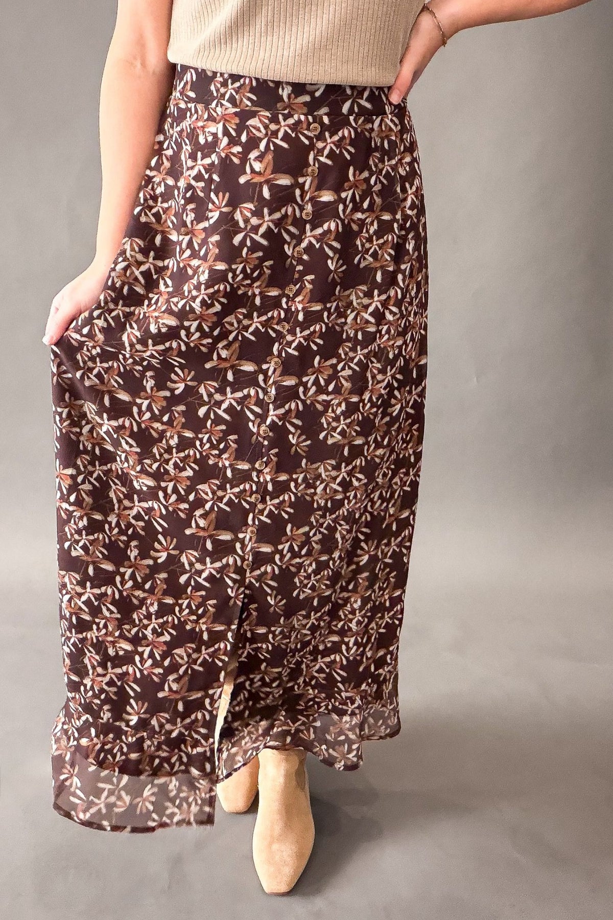 A woman wearing a brown maxi skirt with a beige and white abstract floral pattern, paired with a beige ribbed top and beige ankle boots, standing against a neutral gray studio background.