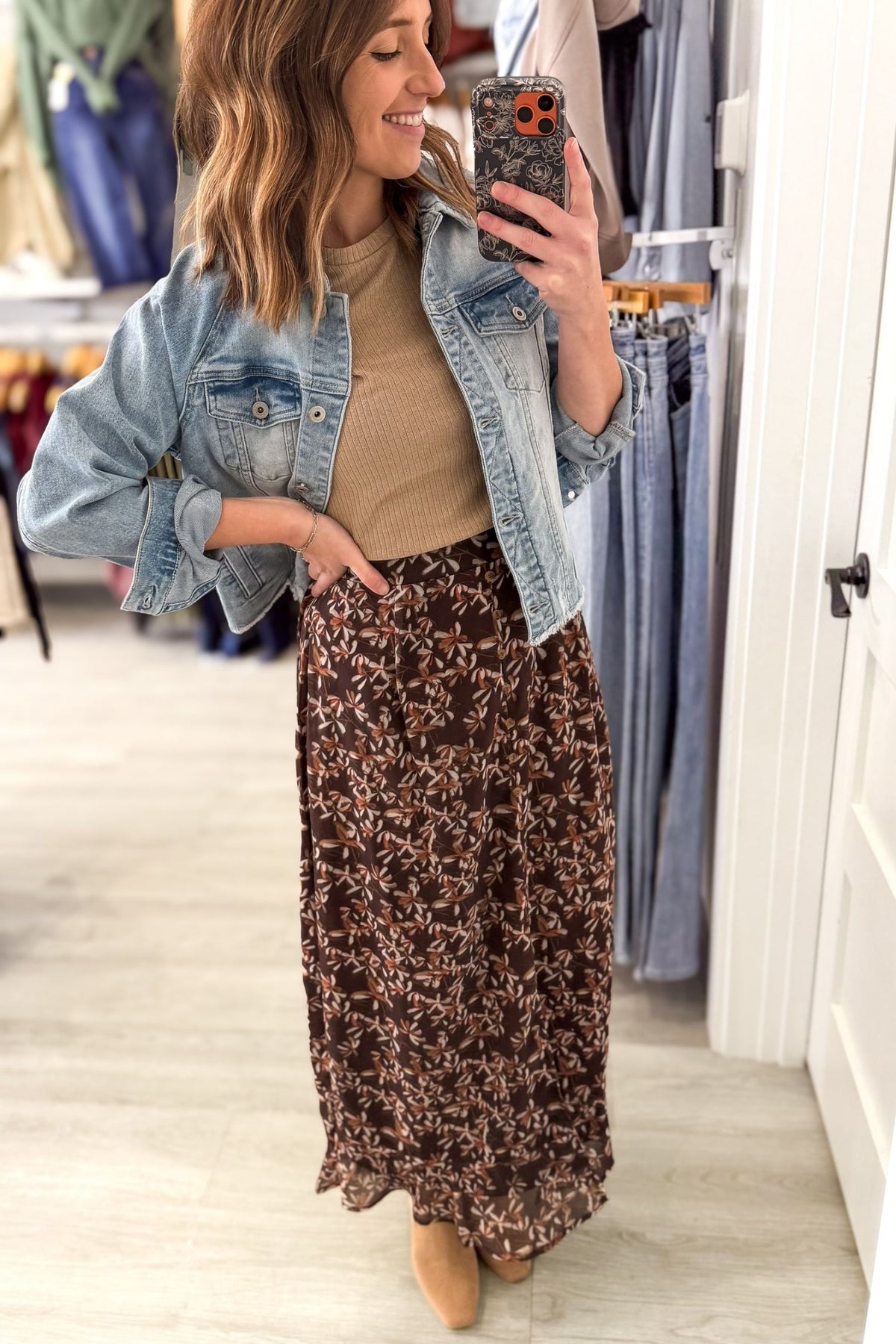 A woman takes a mirror selfie in a clothing store, wearing a light-wash blue denim jacket over a beige ribbed top and a brown floral maxi skirt.
