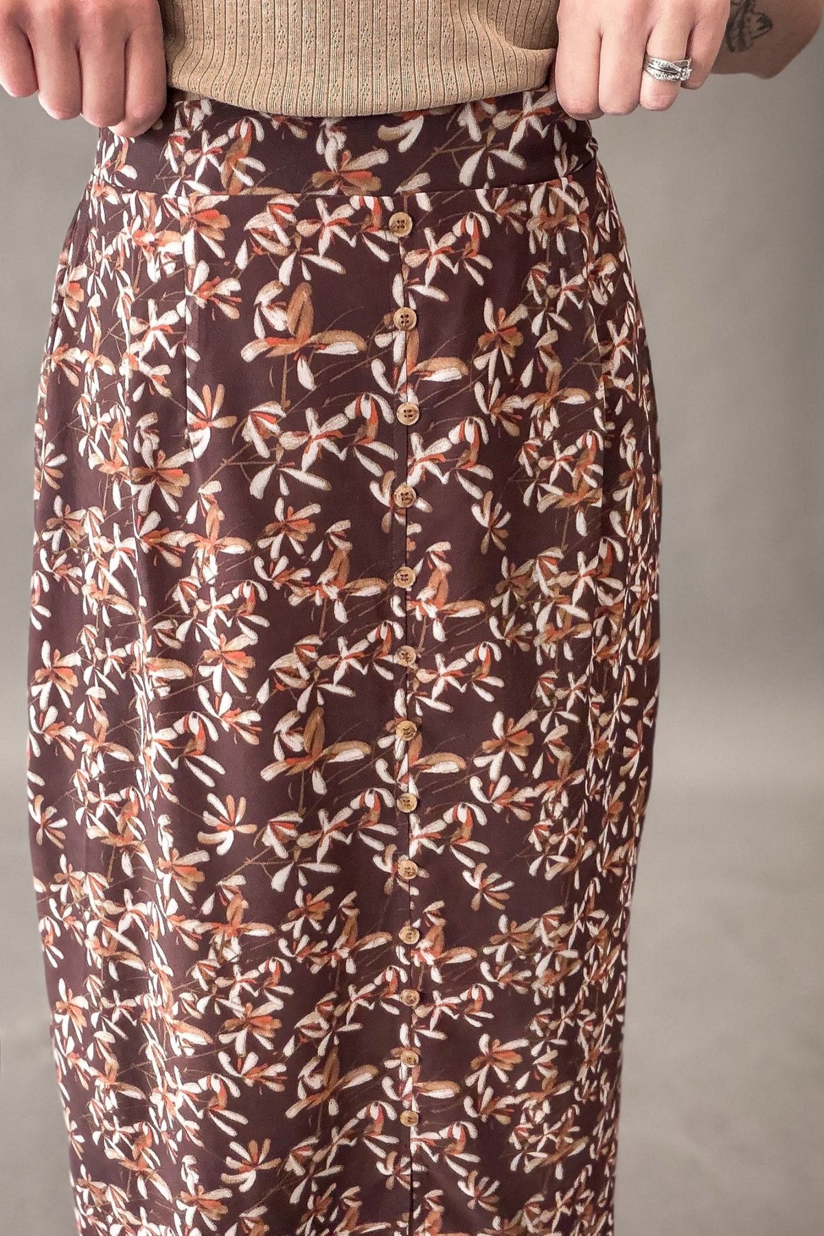 A close-up of a person wearing a brown skirt with an allover floral and butterfly print in beige, white, and rust tones, paired with a beige ribbed top against a neutral gray background.