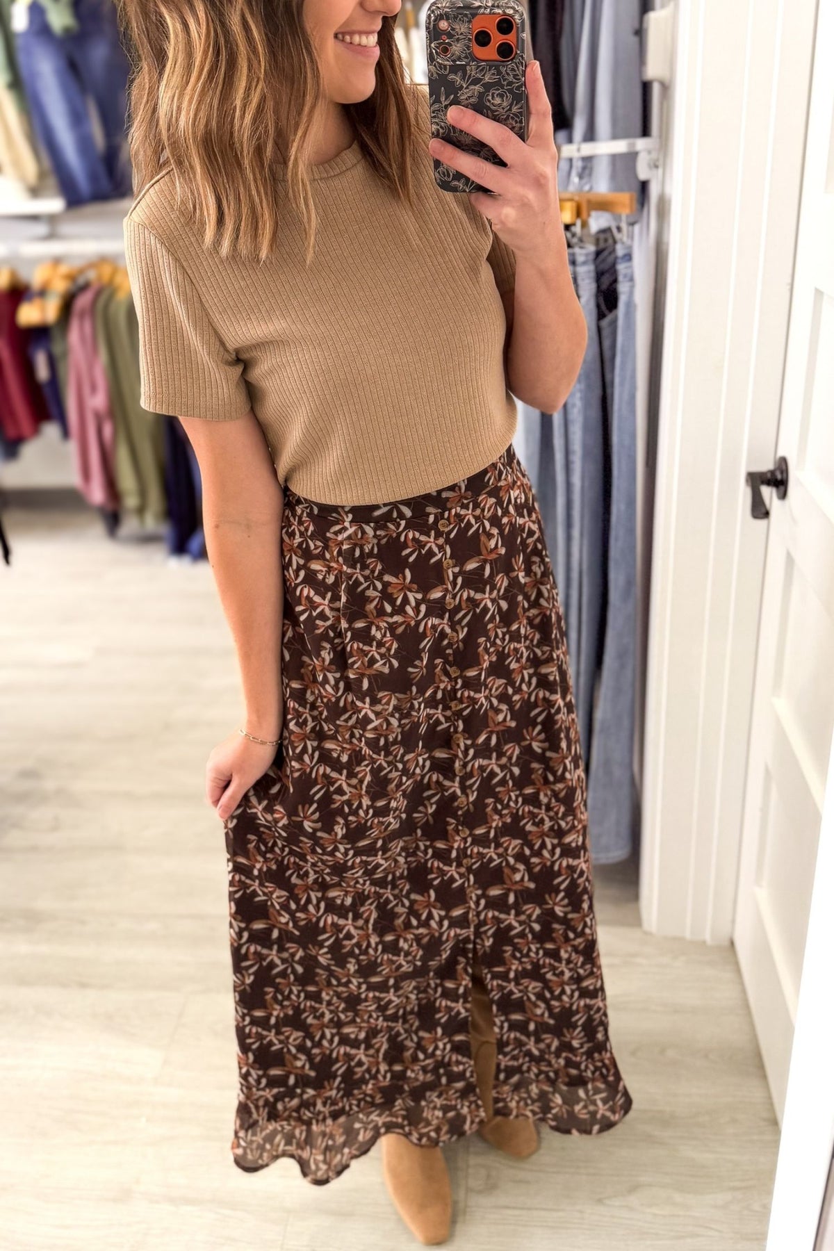 A woman takes a mirror selfie in a clothing store, wearing a beige short-sleeve ribbed knit top and a brown maxi skirt with a beige floral pattern.