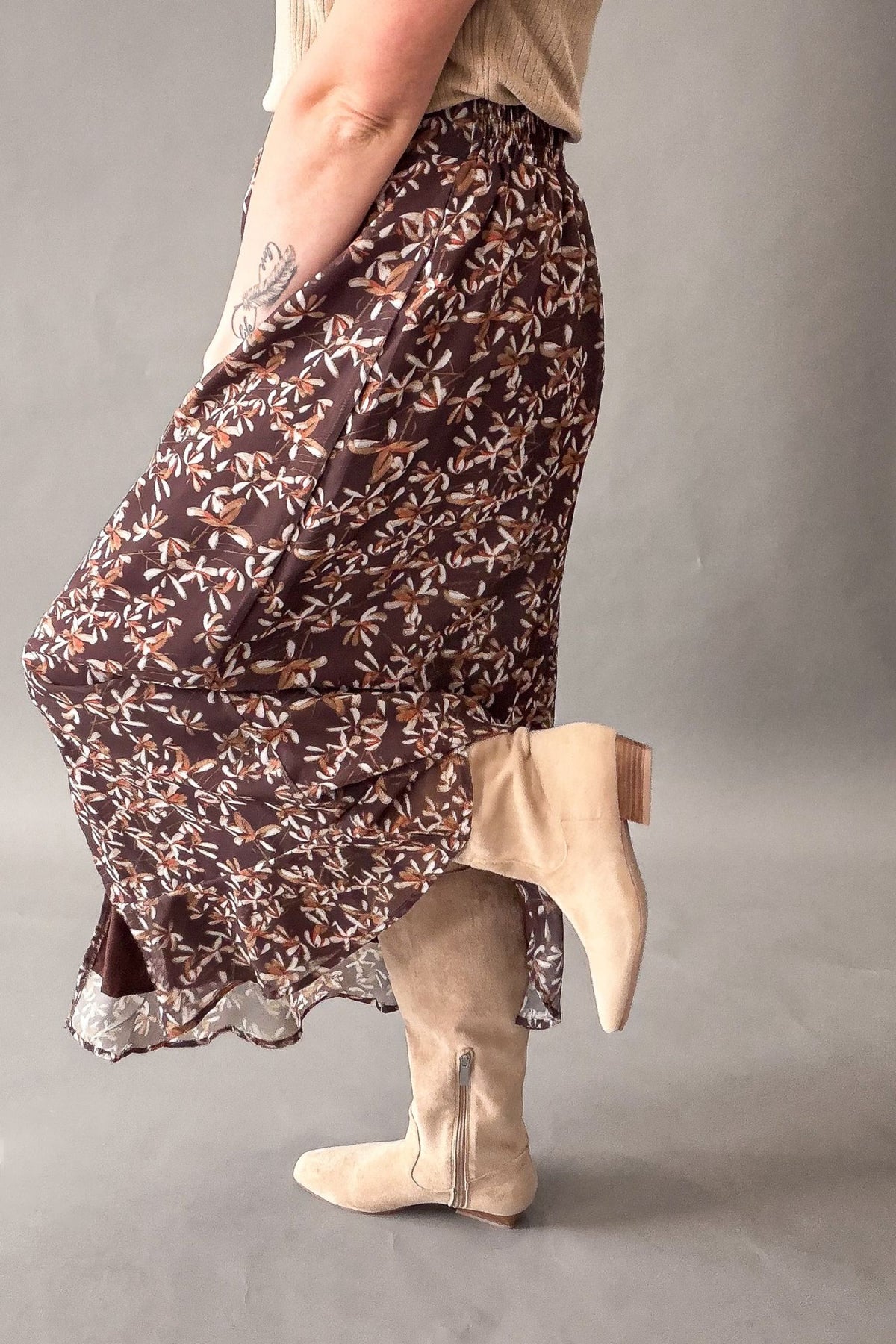 A person wearing a brown maxi skirt with a beige and white floral print, paired with beige suede ankle boots, shown from the waist down against a neutral gray studio background.