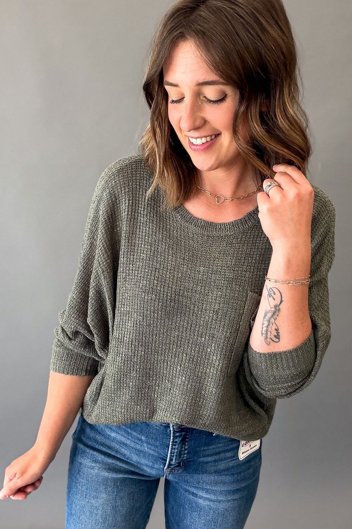 A woman wearing a textured forest green knit sweater and blue jeans, smiling and posing against a solid gray studio background.