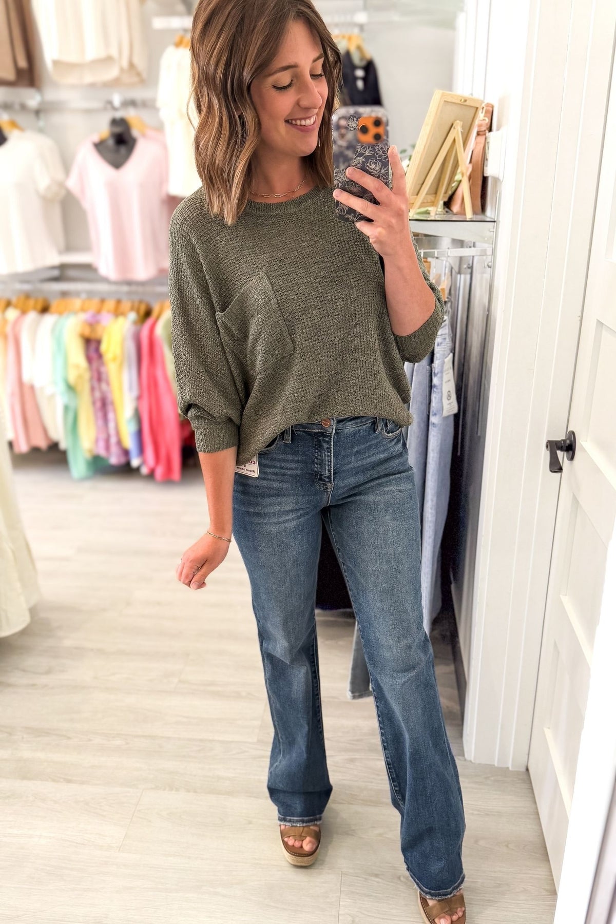 A woman takes a mirror selfie in a clothing boutique wearing an forest green knit 3/4-sleeve top and blue straight jeans.