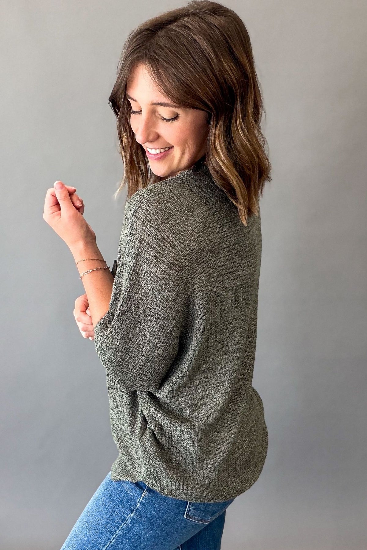 A woman wearing a textured forest green knit top and blue jeans, shown from a side-back angle against a plain gray studio background.