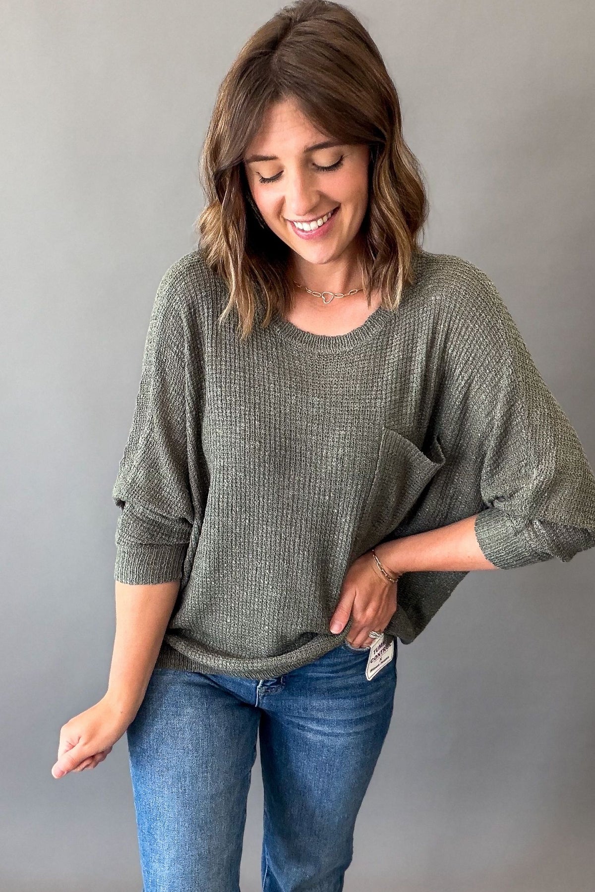 A woman wearing a textured forest green knit top with three-quarter sleeves and blue jeans, standing against a plain gray background.