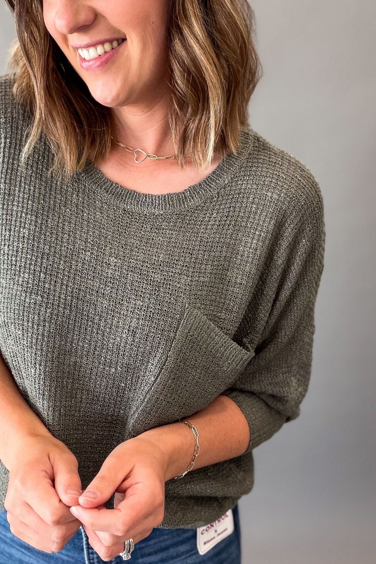 A woman wearing a textured forest green knit sweater with a chest pocket, paired with blue jeans, shown in a close-up shot against a plain gray background.