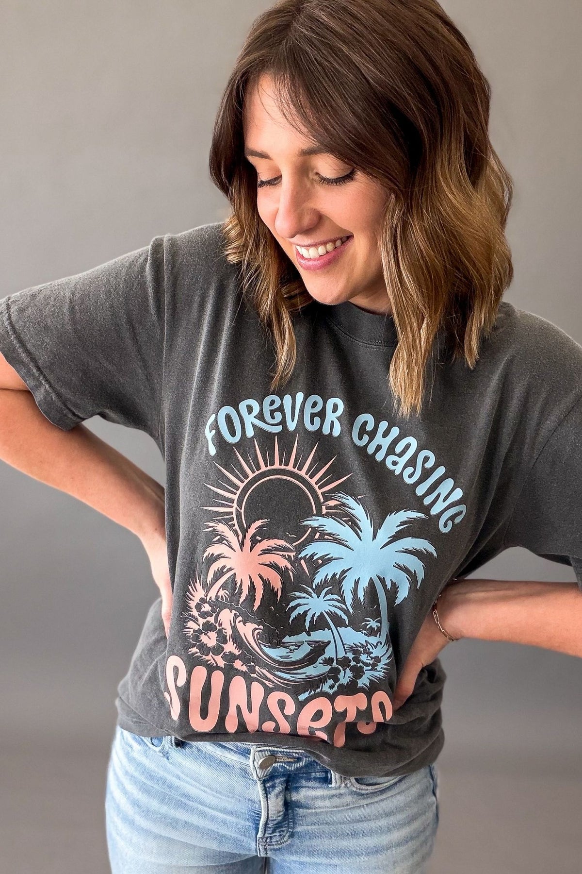Woman wearing a pepper colored Forever Chasing Sunsets graphic tee with palm tree sunset design and casual denim outfit