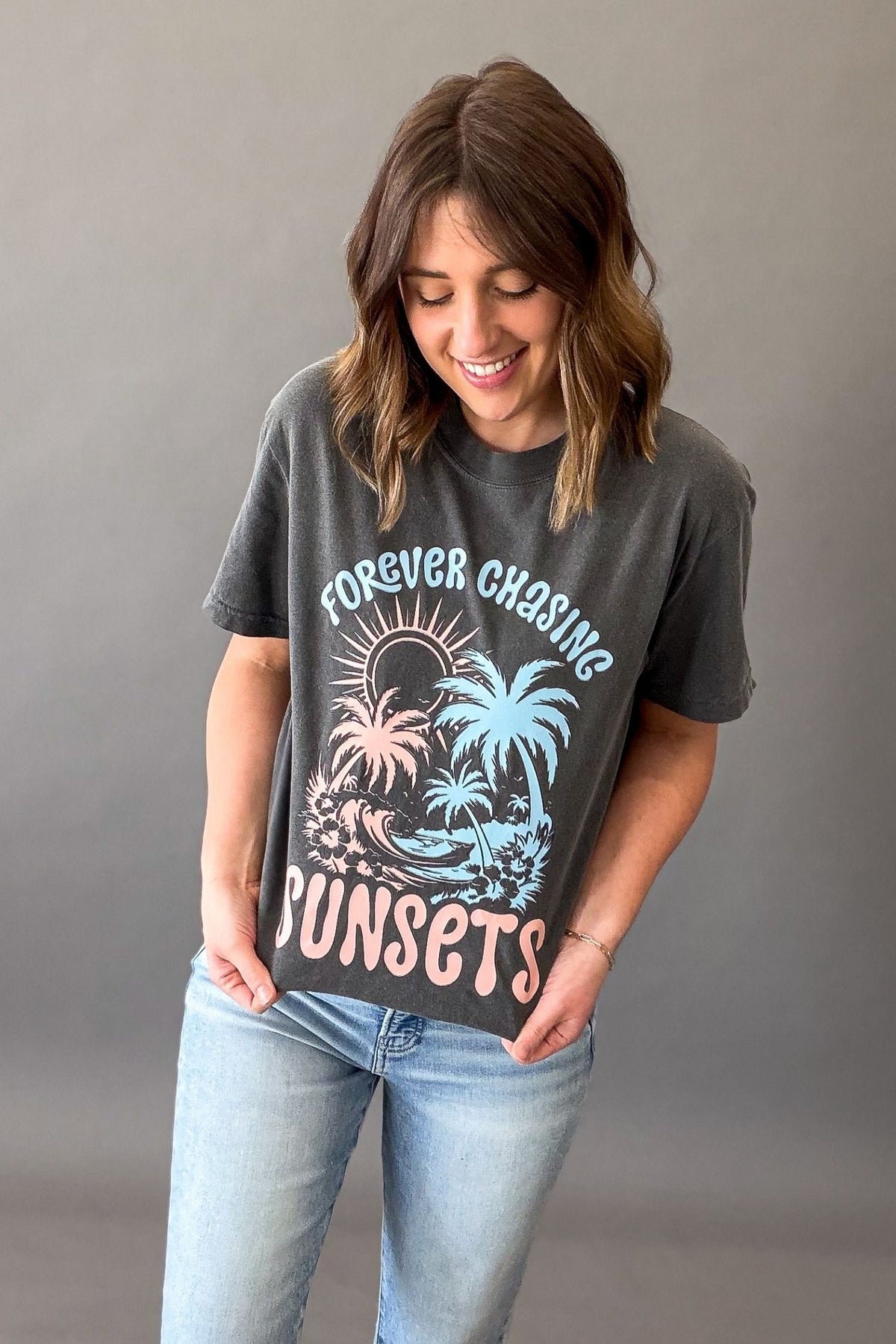 Woman wearing a pepper colored Forever Chasing Sunsets graphic tee with palm tree sunset design and casual denim outfit