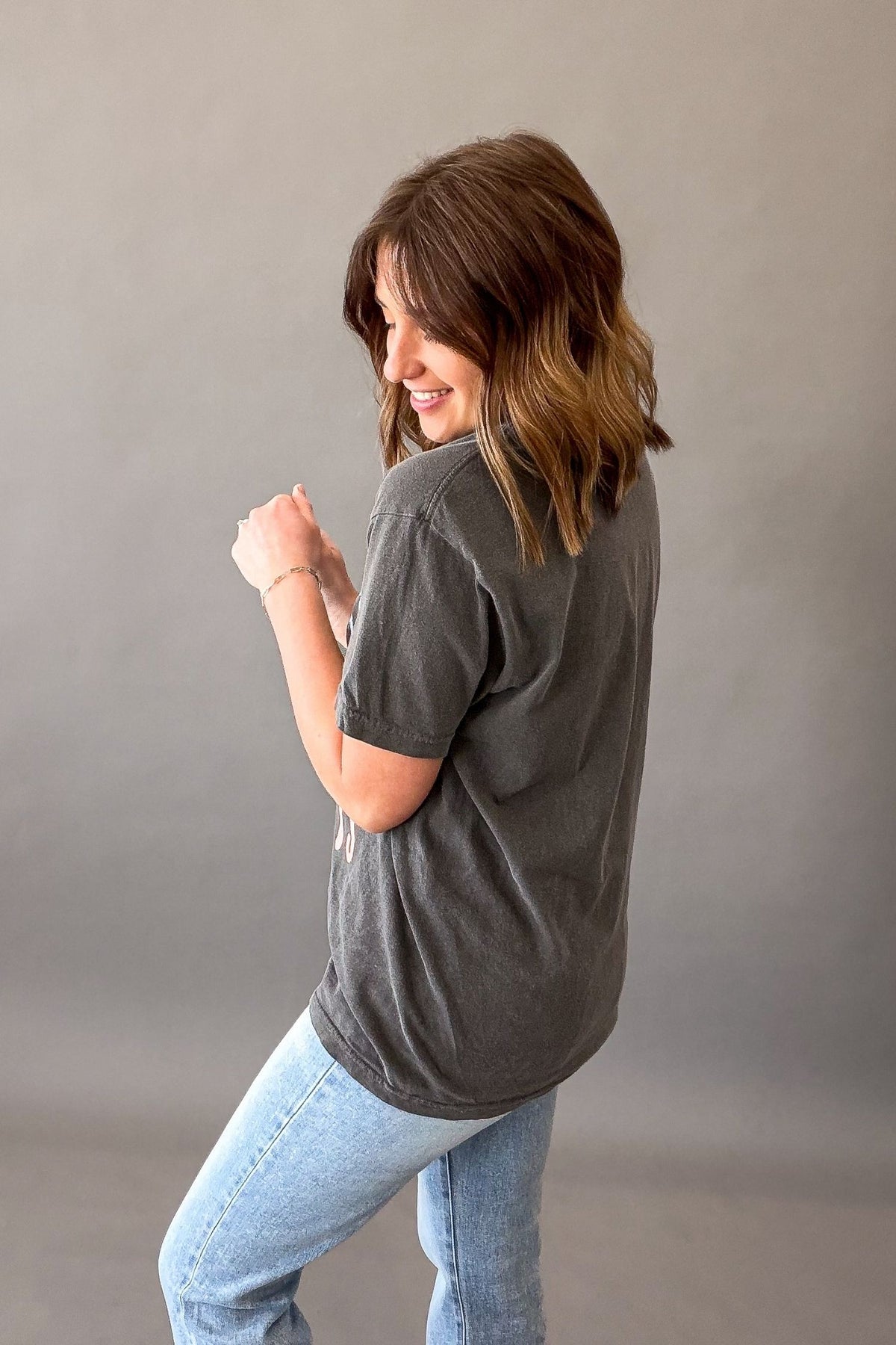 Woman wearing a pepper colored Forever Chasing Sunsets graphic tee with palm tree sunset design and casual denim outfit showing back details