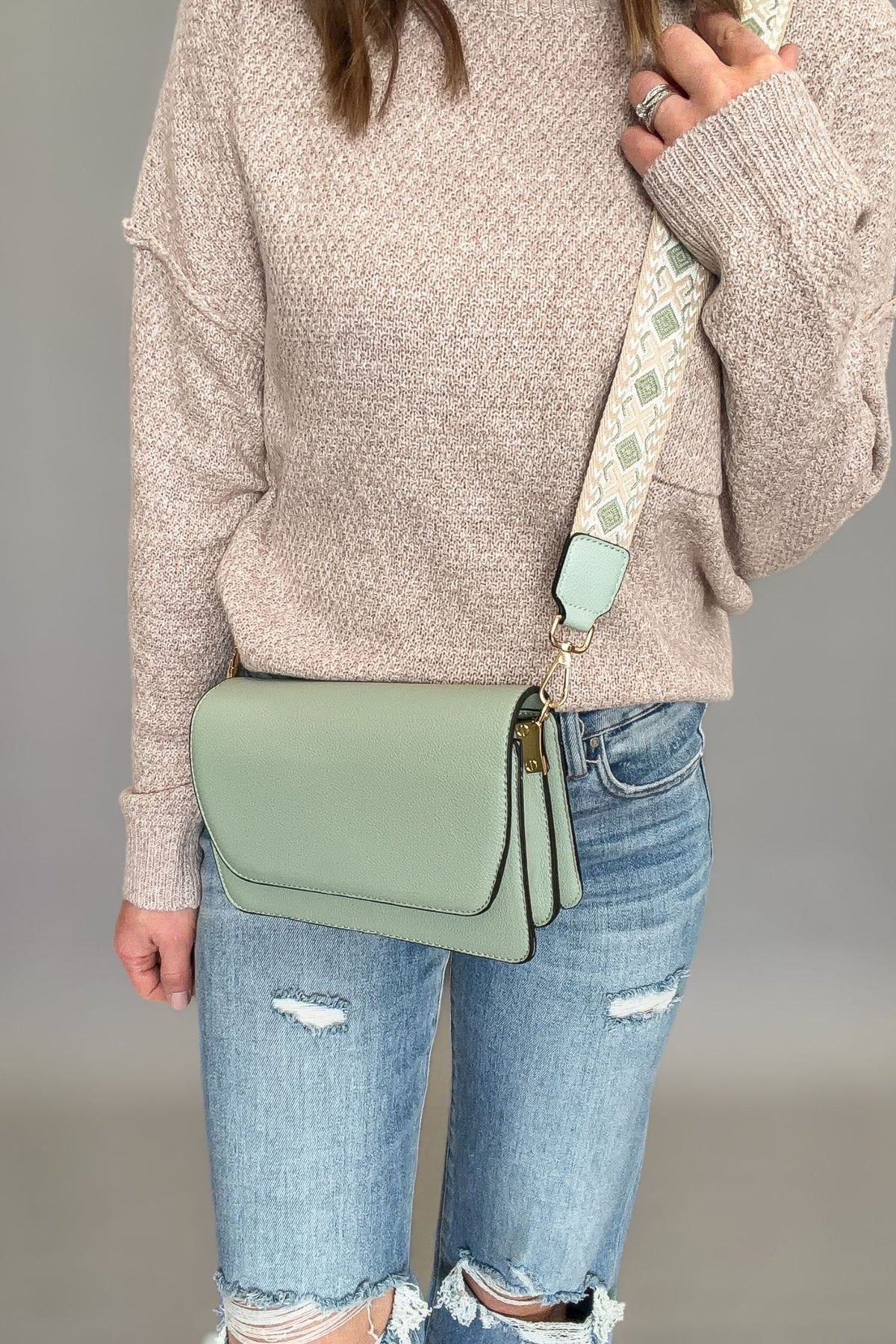 Person wearing a beige sweater and blue jeans, holding a green crossbody bag with a patterned strap.