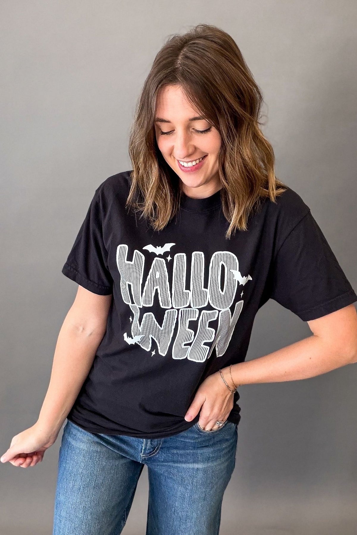 A woman wearing a black graphic t-shirt with 'HALLOWEEN' printed in distressed white and gray lettering, paired with blue jeans, posing against a solid gray studio background.