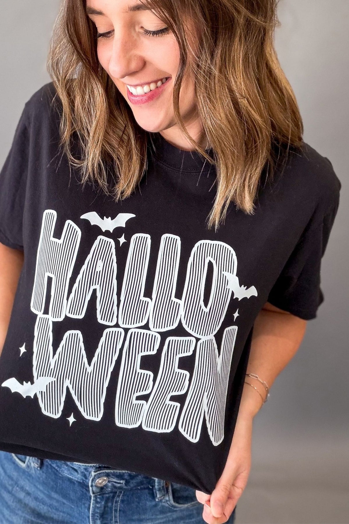 A woman wearing a black graphic t-shirt with 'HALLOWEEN' printed in white striped letters, paired with blue jeans, against a plain gray background.