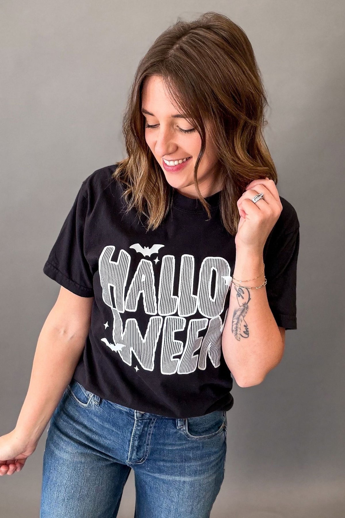 A woman wearing a black graphic t-shirt with 'HALLOWEEN' printed in white and gray, paired with blue jeans, posing against a neutral gray background.