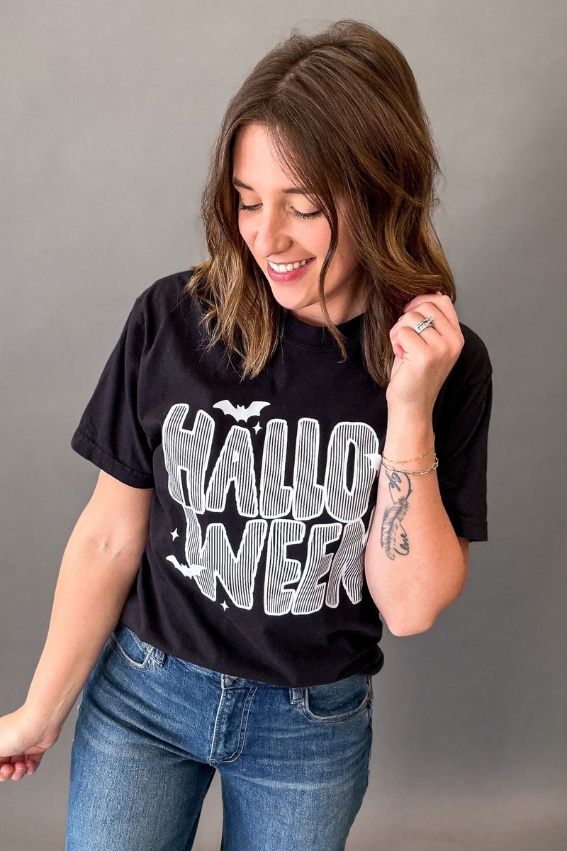A woman wearing a black graphic t-shirt with 'HALLOWEEN' printed in white and gray, paired with blue jeans, posing against a neutral gray background.
