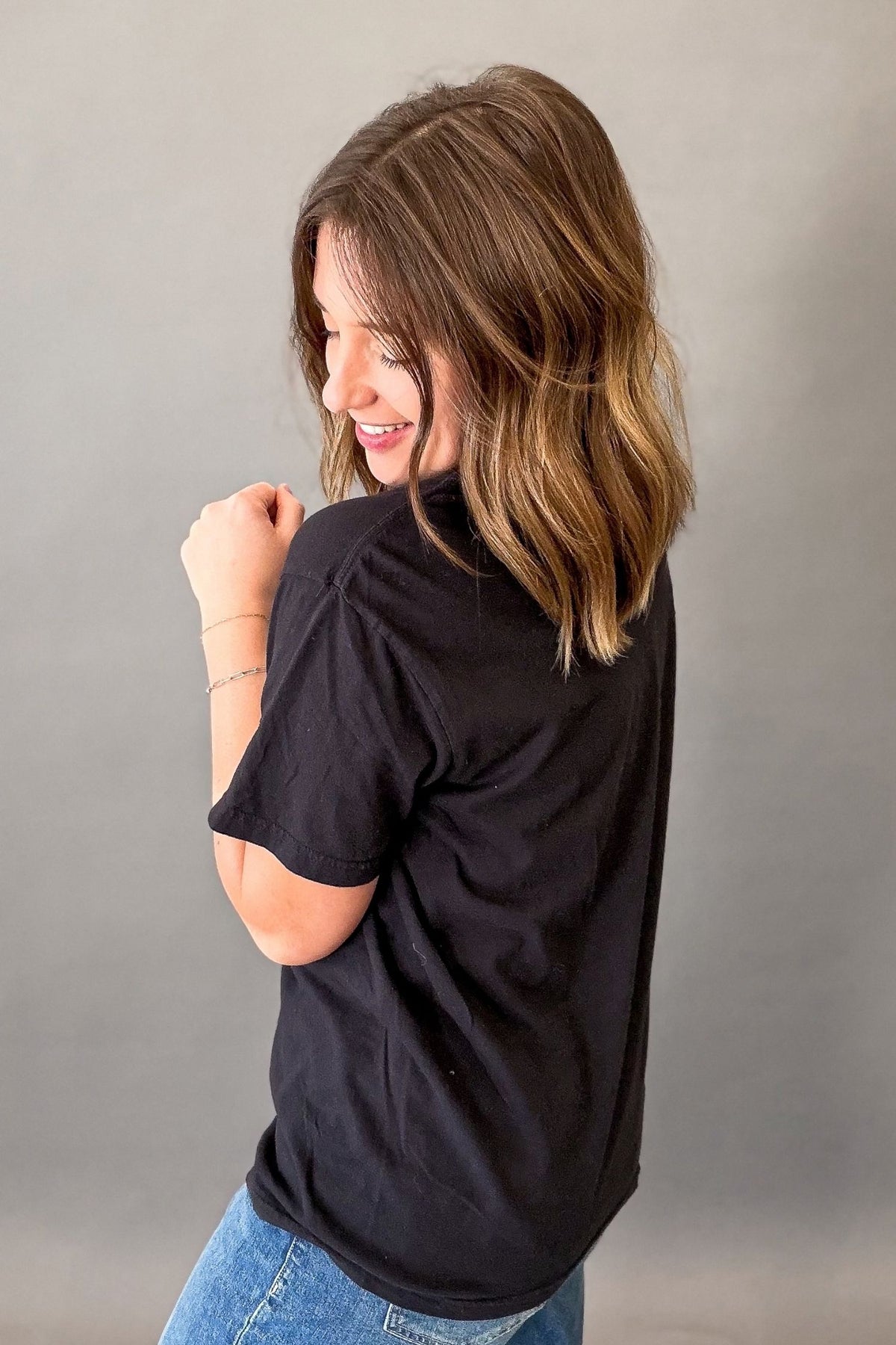A woman with shoulder-length brown hair is shown from the back wearing a black short-sleeve t-shirt and blue jeans against a plain gray studio background.
