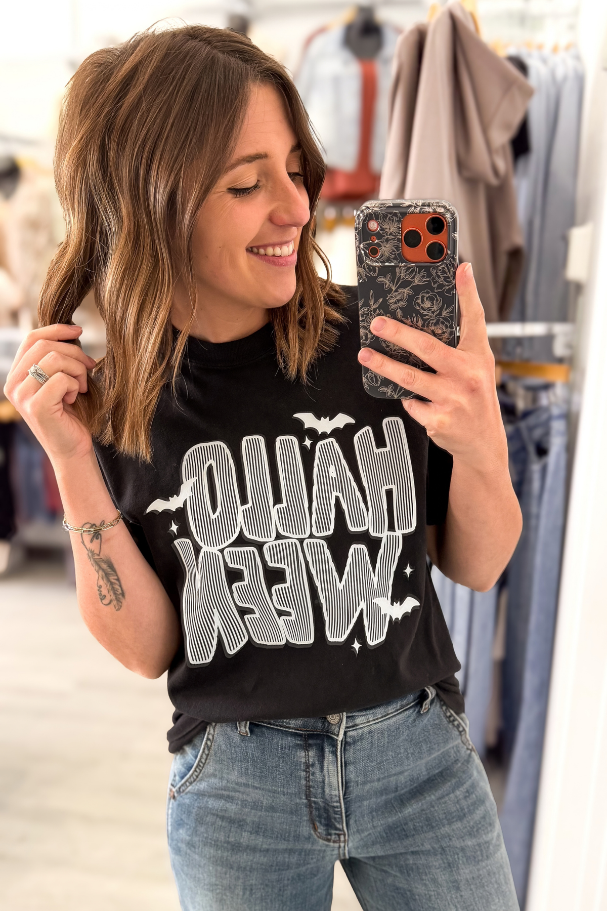 A woman taking a mirror selfie in a clothing store, wearing a black graphic t-shirt with 'HALLOWEEN' written in distressed white letters with bat silhouettes.
