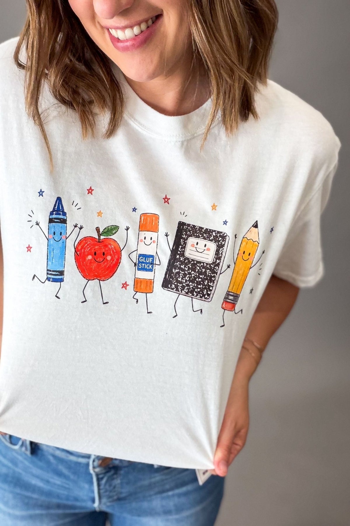 A woman wearing a white graphic t-shirt featuring cute, smiling school supplies: a blue crayon, a red apple, a glue stick, a notebook, and a yellow pencil. She is also wearing blue jeans against a neutral gray background.