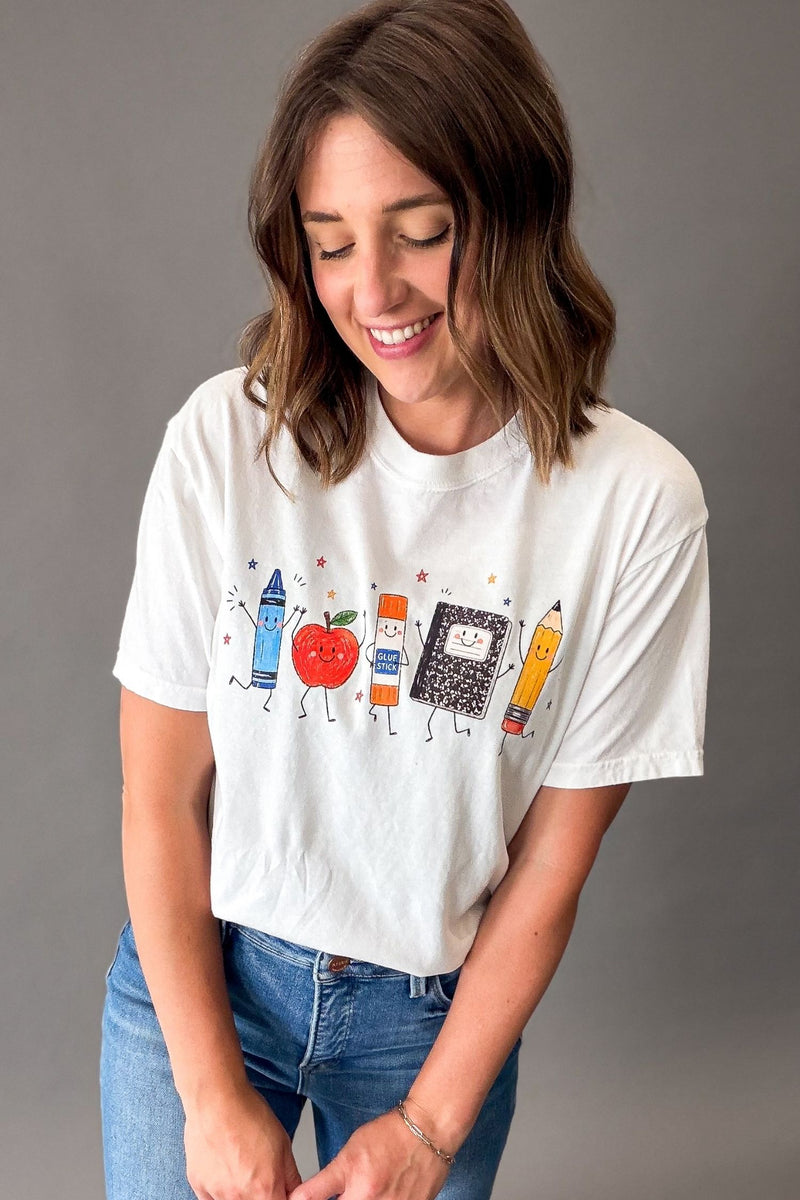 A woman wearing a white graphic t-shirt with a colorful school supply design, paired with blue jeans, against a solid gray studio background.