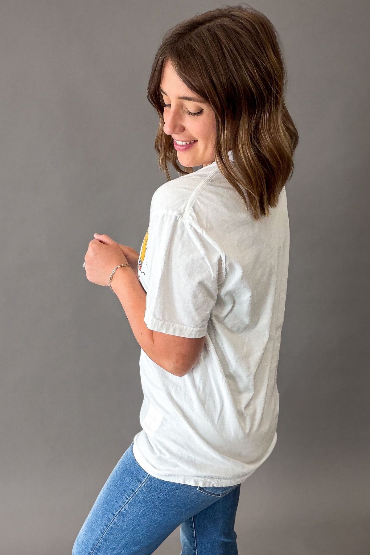 A woman with shoulder-length brown hair is shown from the back wearing a white short-sleeve t-shirt and blue jeans against a solid gray studio background.