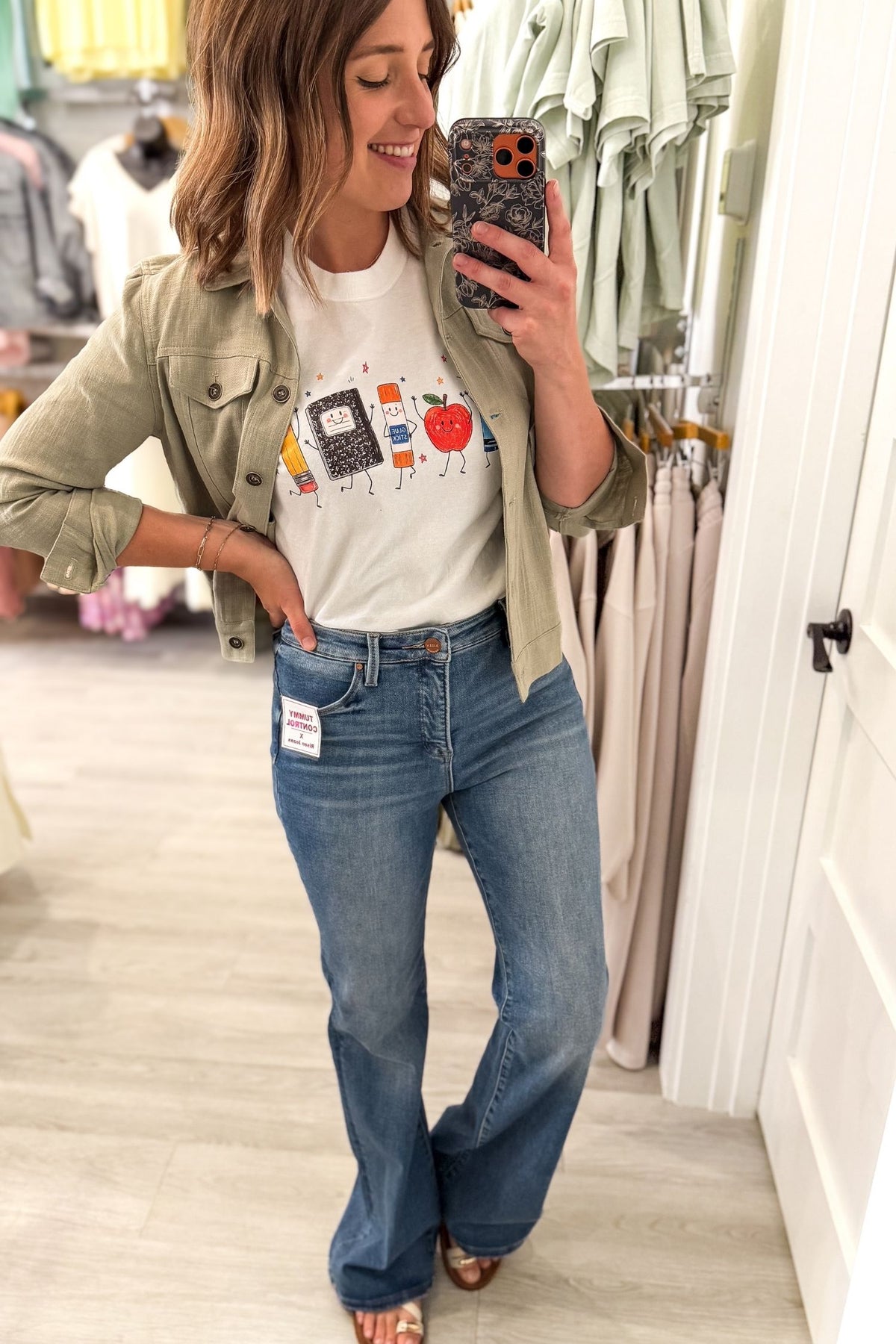 A woman takes a mirror selfie in a clothing store wearing a white graphic t-shirt, an olive green button-up jacket, and blue flare jeans.