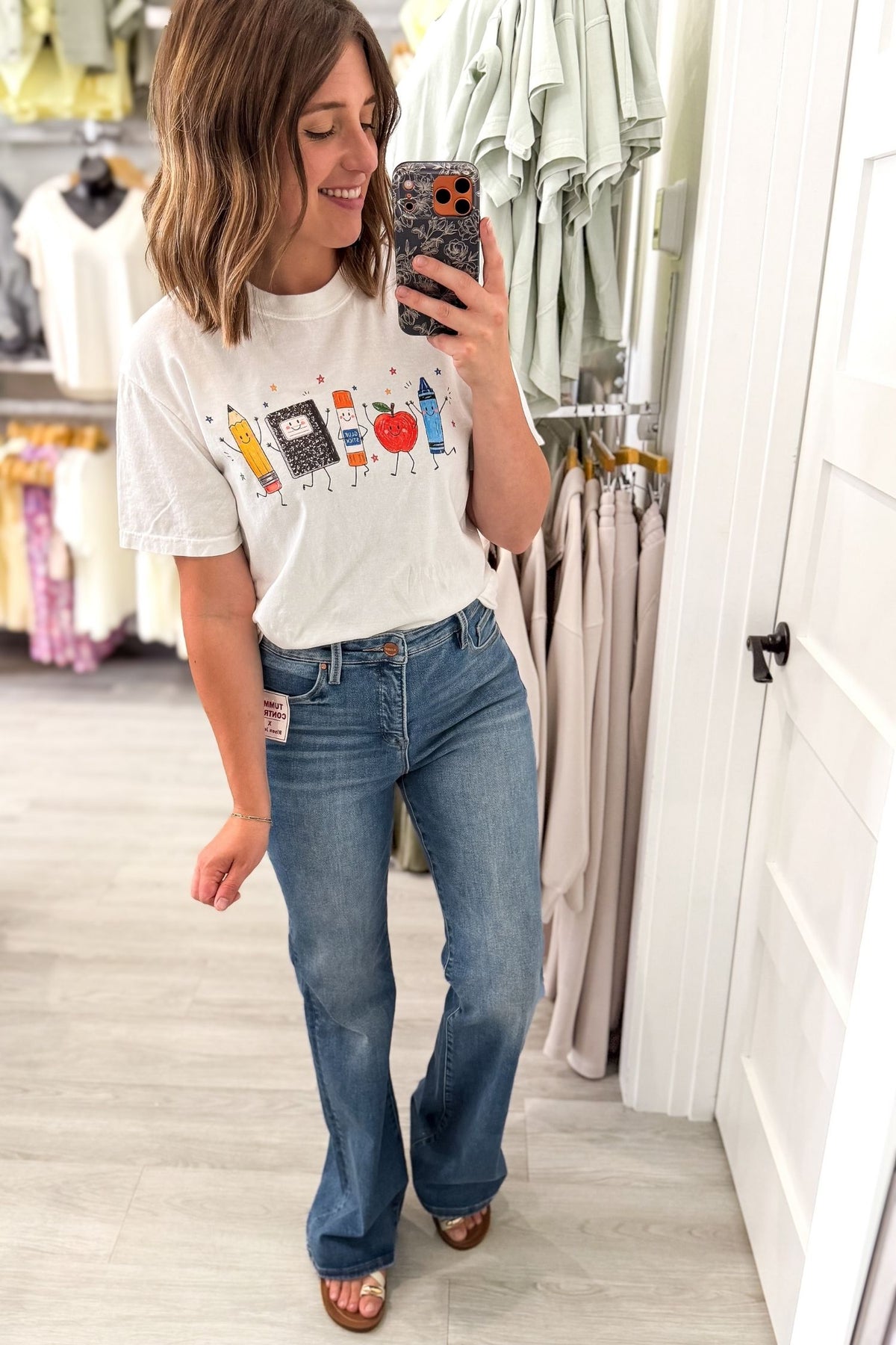A woman takes a mirror selfie in a clothing store wearing a white graphic t-shirt with colorful illustrations, blue flare jeans, and brown sandals.