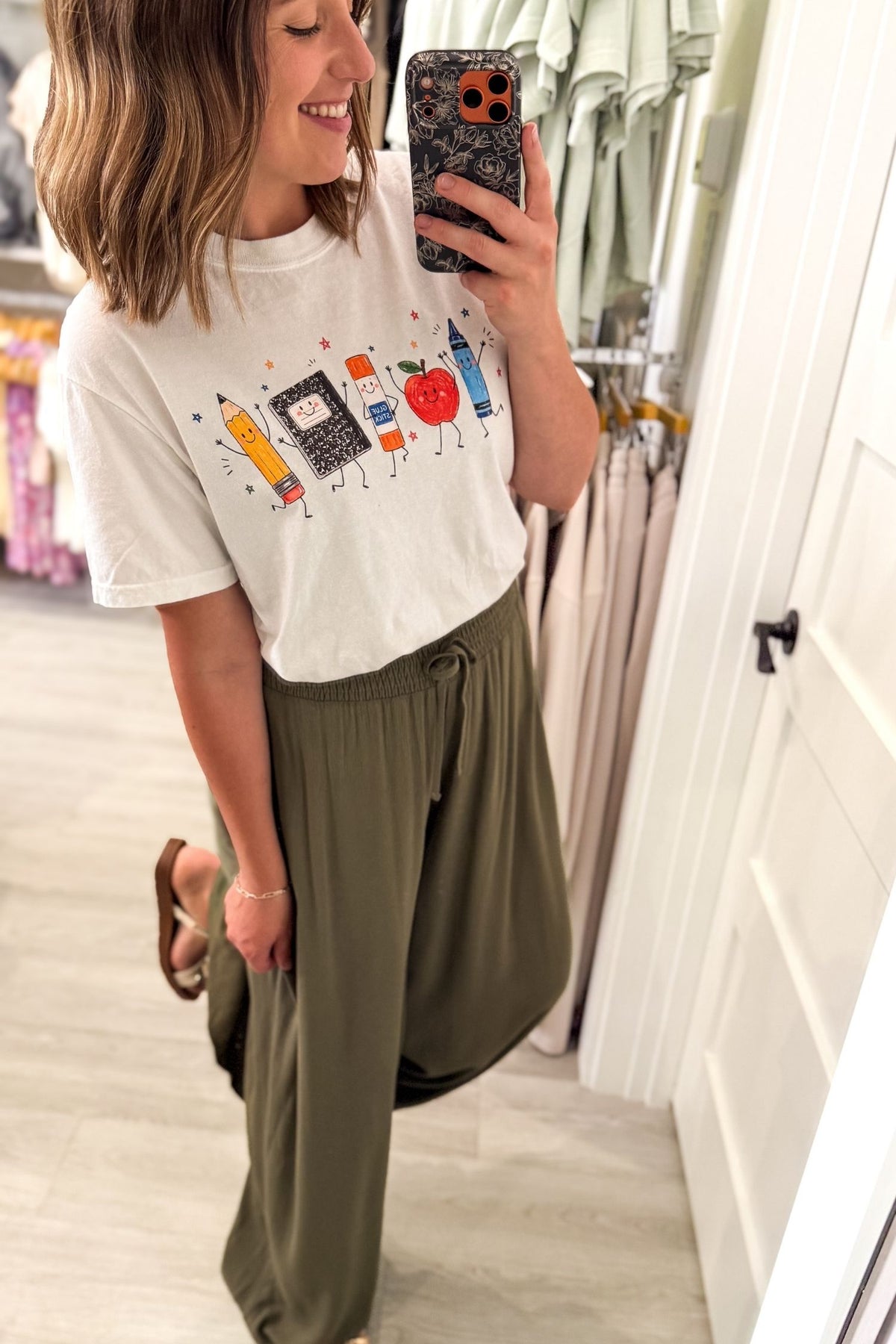 A woman takes a mirror selfie in a boutique showing a white graphic t-shirt with colorful paintbrushes and a red apple, paired with olive green wide-leg drawstring pants.