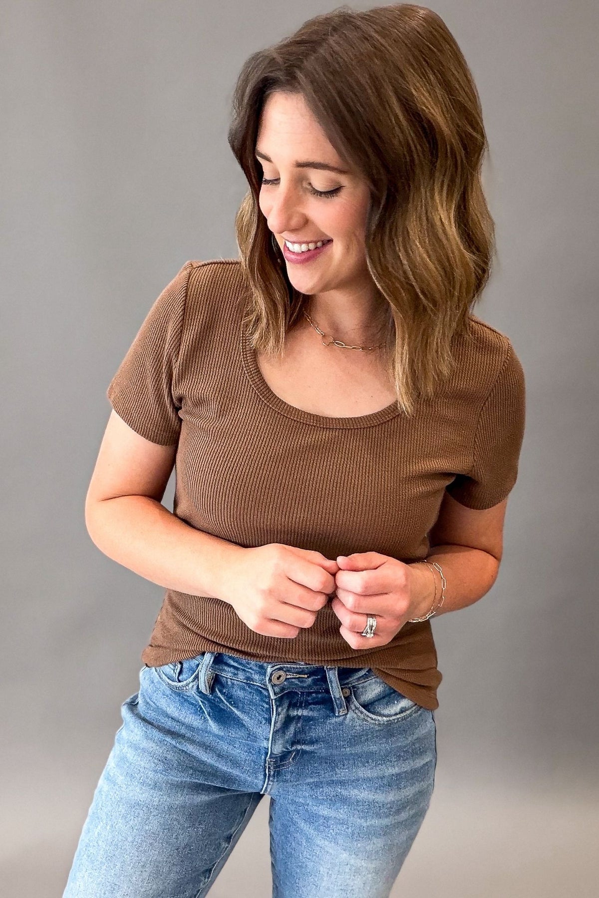 A woman wearing a hazelnut ribbed short-sleeve scoop neck t-shirt and blue jeans, standing against a neutral gray studio background.