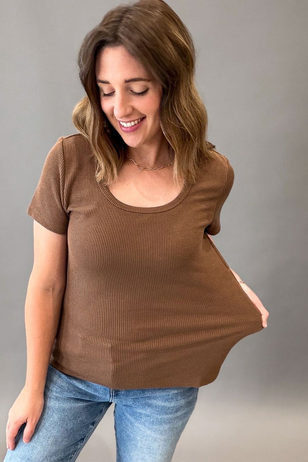 A woman wearing a hazelnut ribbed short-sleeve scoop-neck maternity top and blue jeans, holding out her side of the shirt to show its stretch, against a solid gray background.