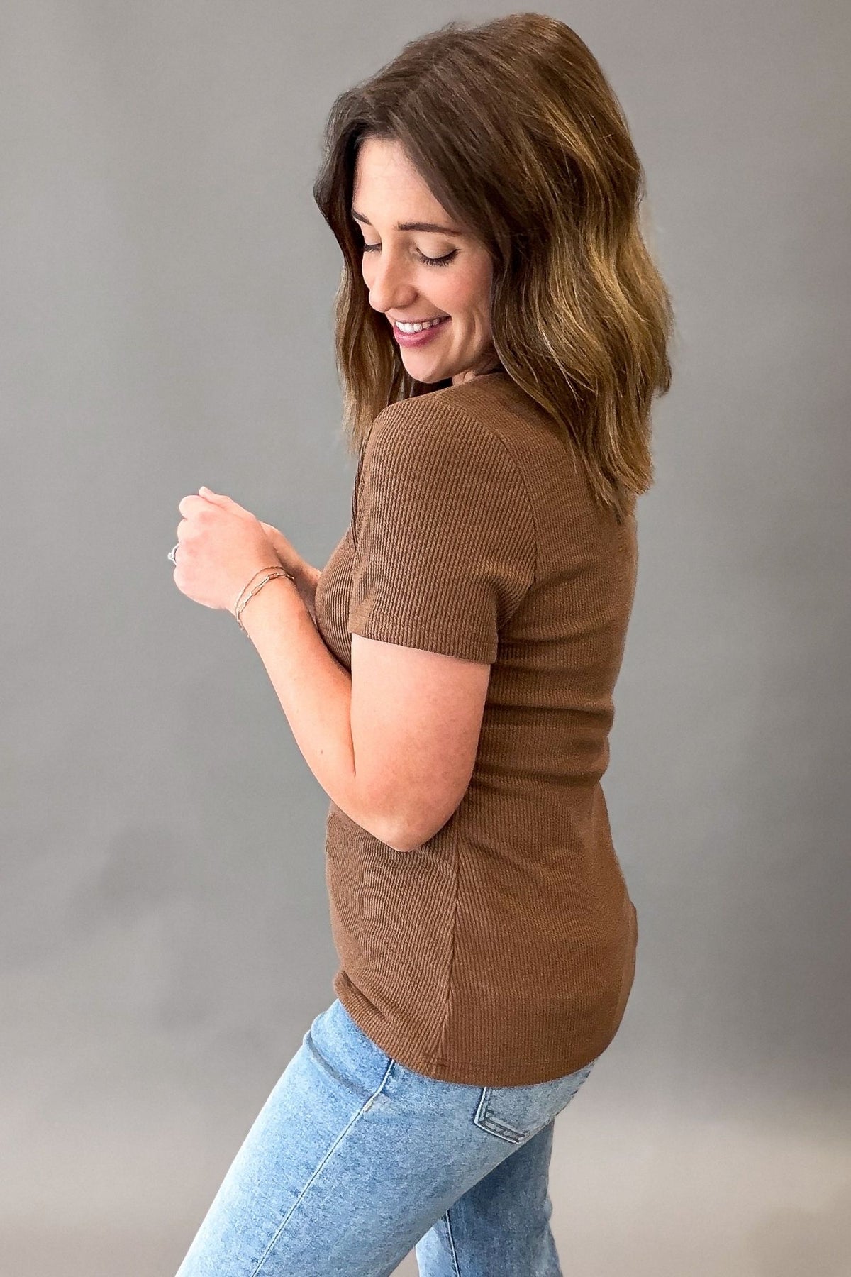 A woman wearing a hazlenut short-sleeve ribbed knit top and light blue jeans, shown from a side-back angle against a plain gray studio background.