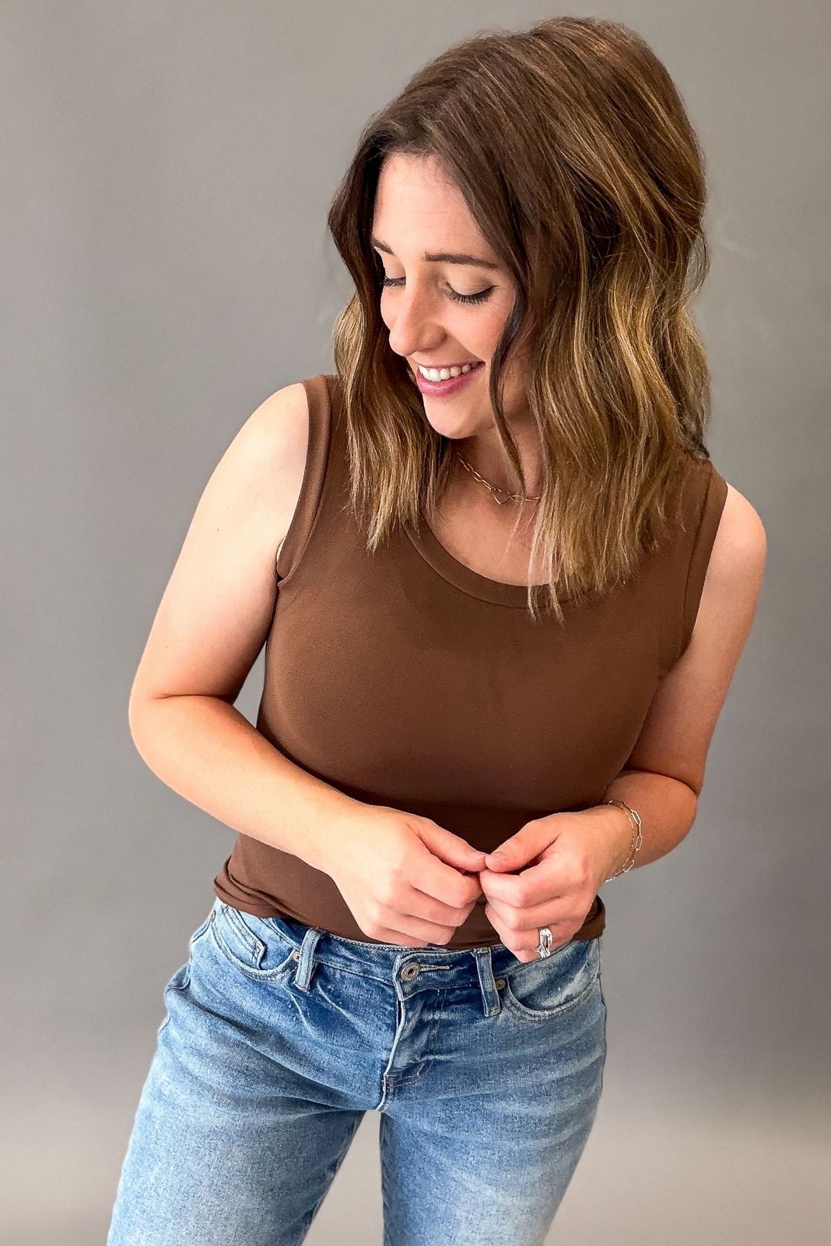 A woman wearing a brown sleeveless tank top and blue jeans, standing against a plain gray studio background.