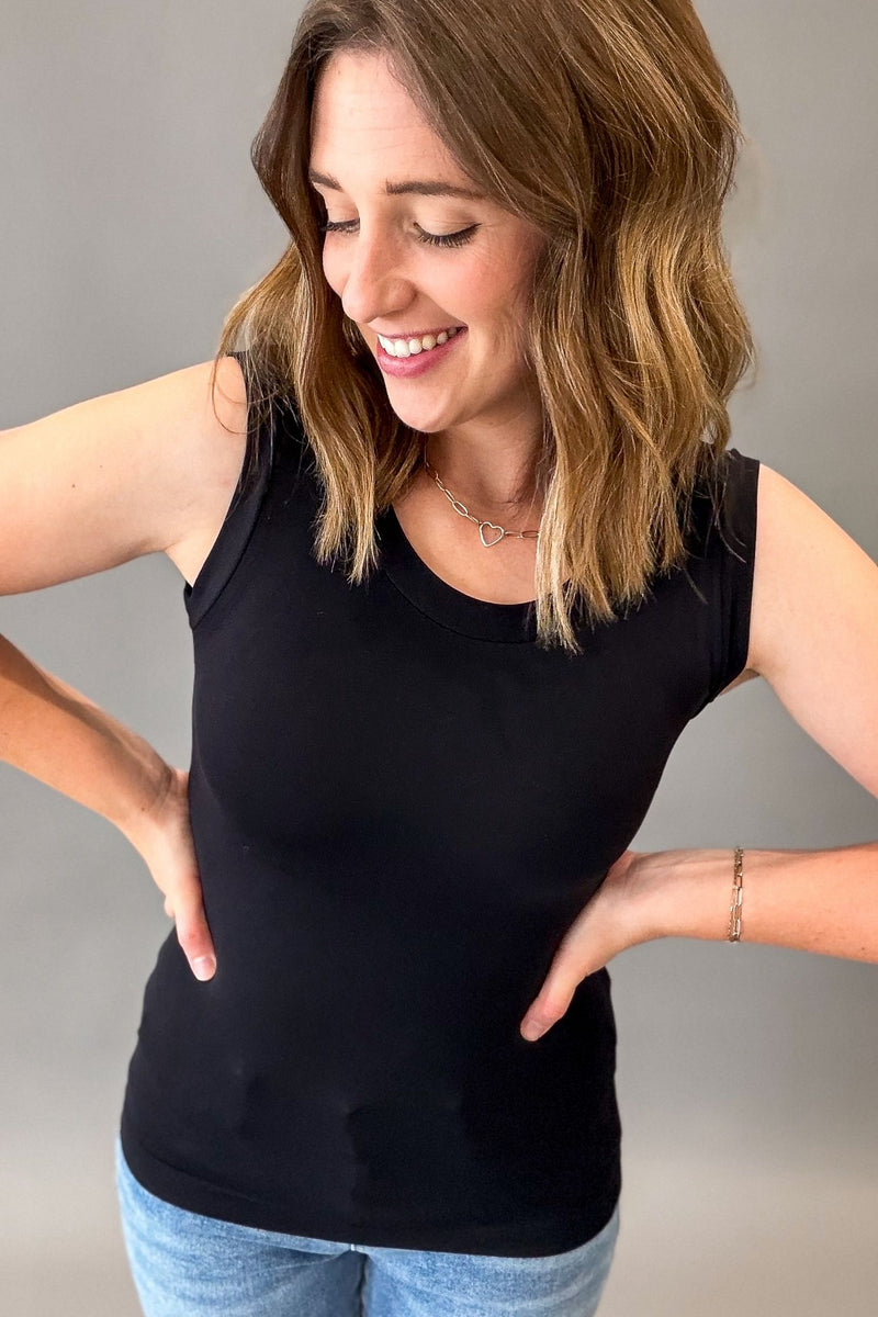 A woman wearing a black sleeveless tank top and light blue jeans, posing with her hands on her hips against a plain gray studio background.