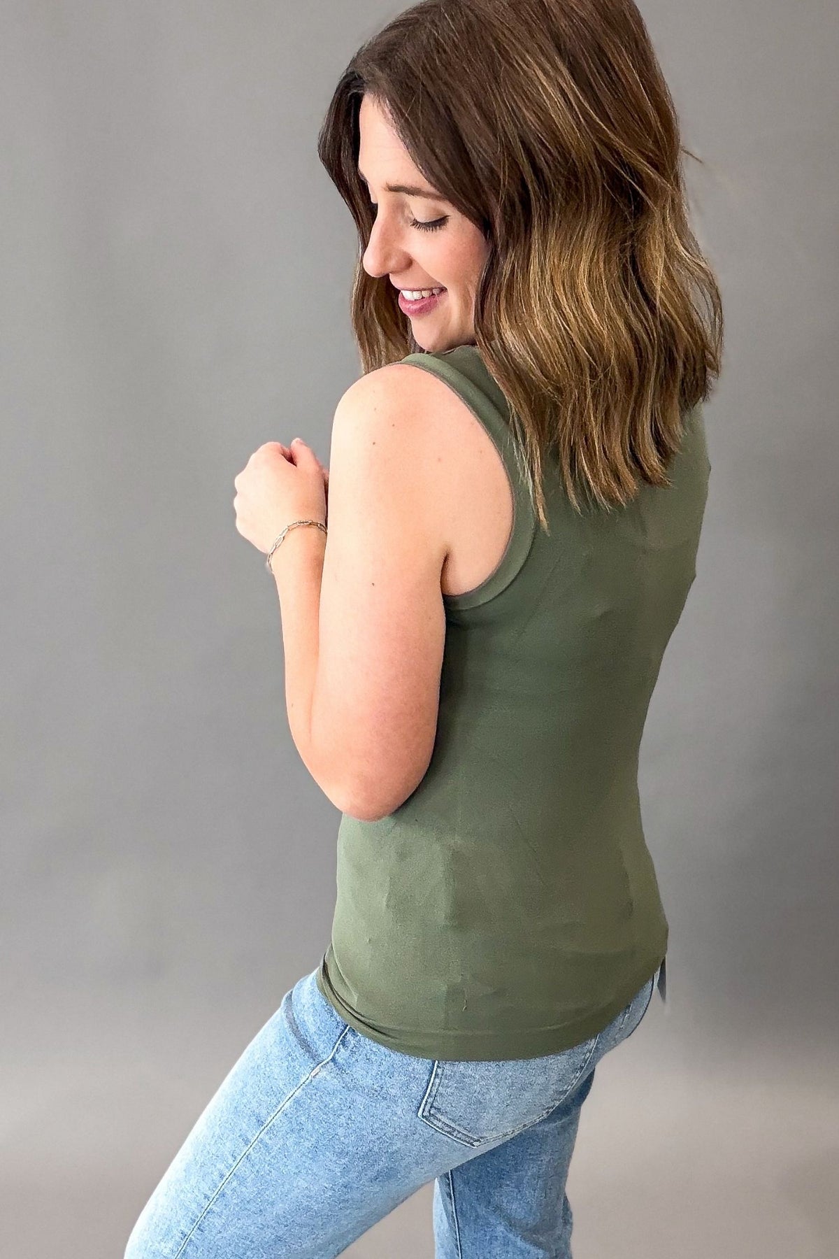 A woman wearing an olive green sleeveless tank top and light blue jeans, shown from a side-back angle against a plain gray studio background.