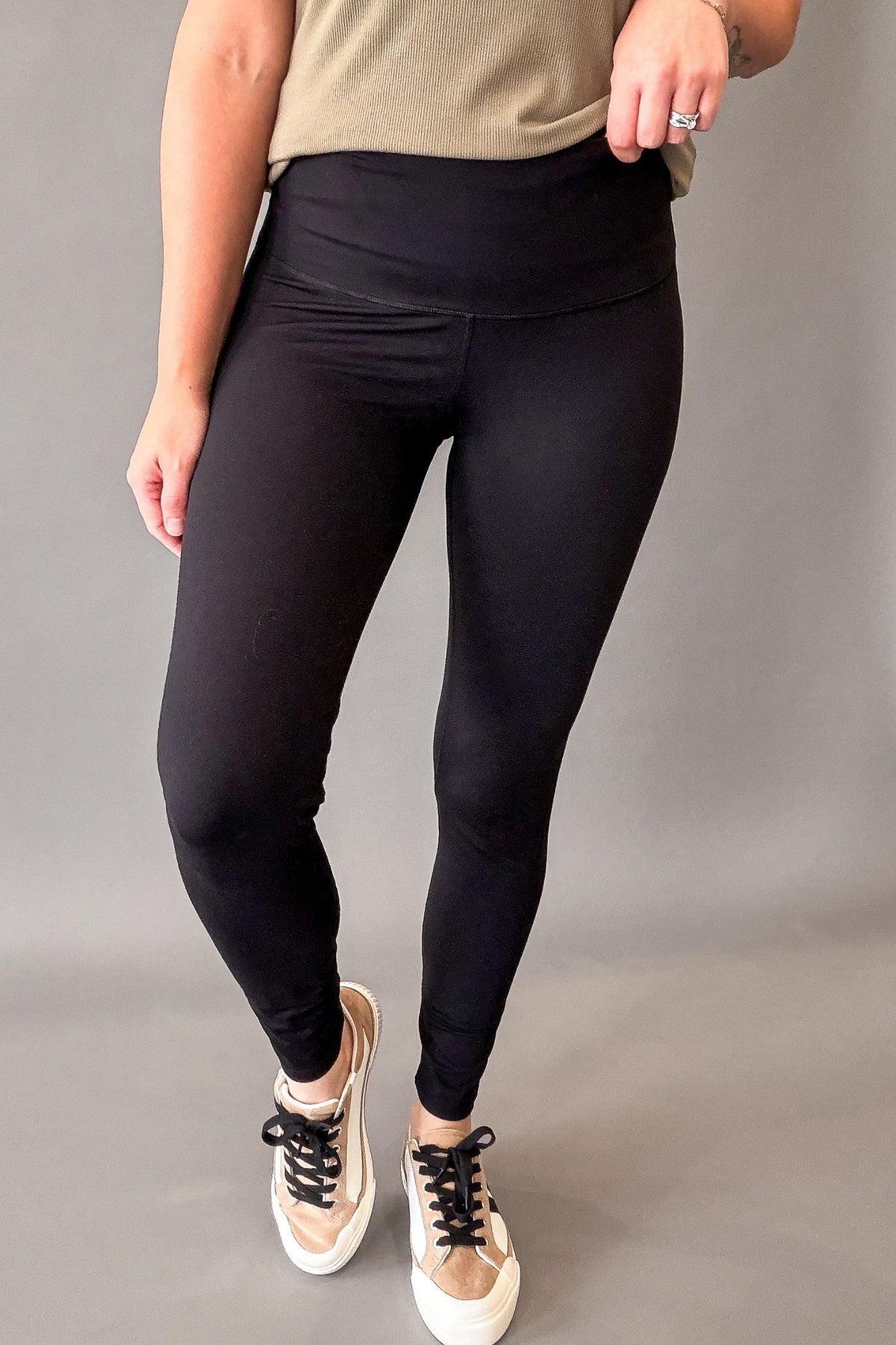 A person modeling black high-waisted leggings with a beige top and beige sneakers against a plain gray studio background.