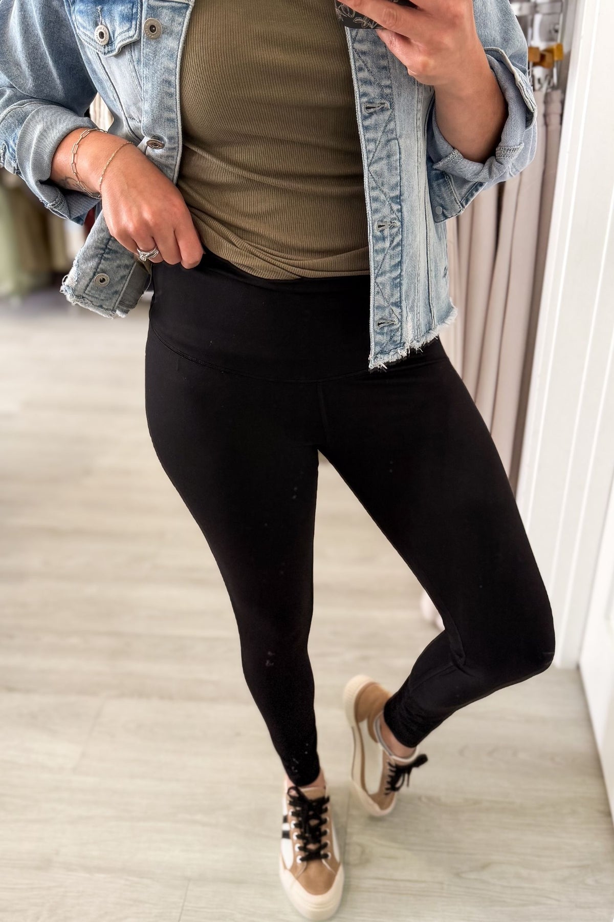 A person wearing black leggings, an olive green ribbed tank top, a light-wash denim jacket, and beige sneakers, shown in a mirror selfie in an indoor room with light wood flooring.