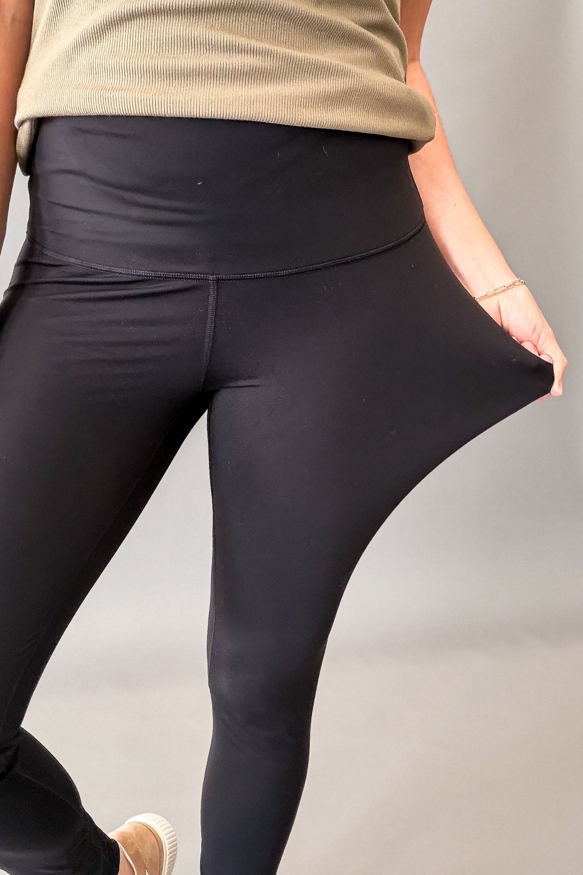Black high-waisted leggings being stretched by a model's hand, with a beige ribbed tank top partially visible.