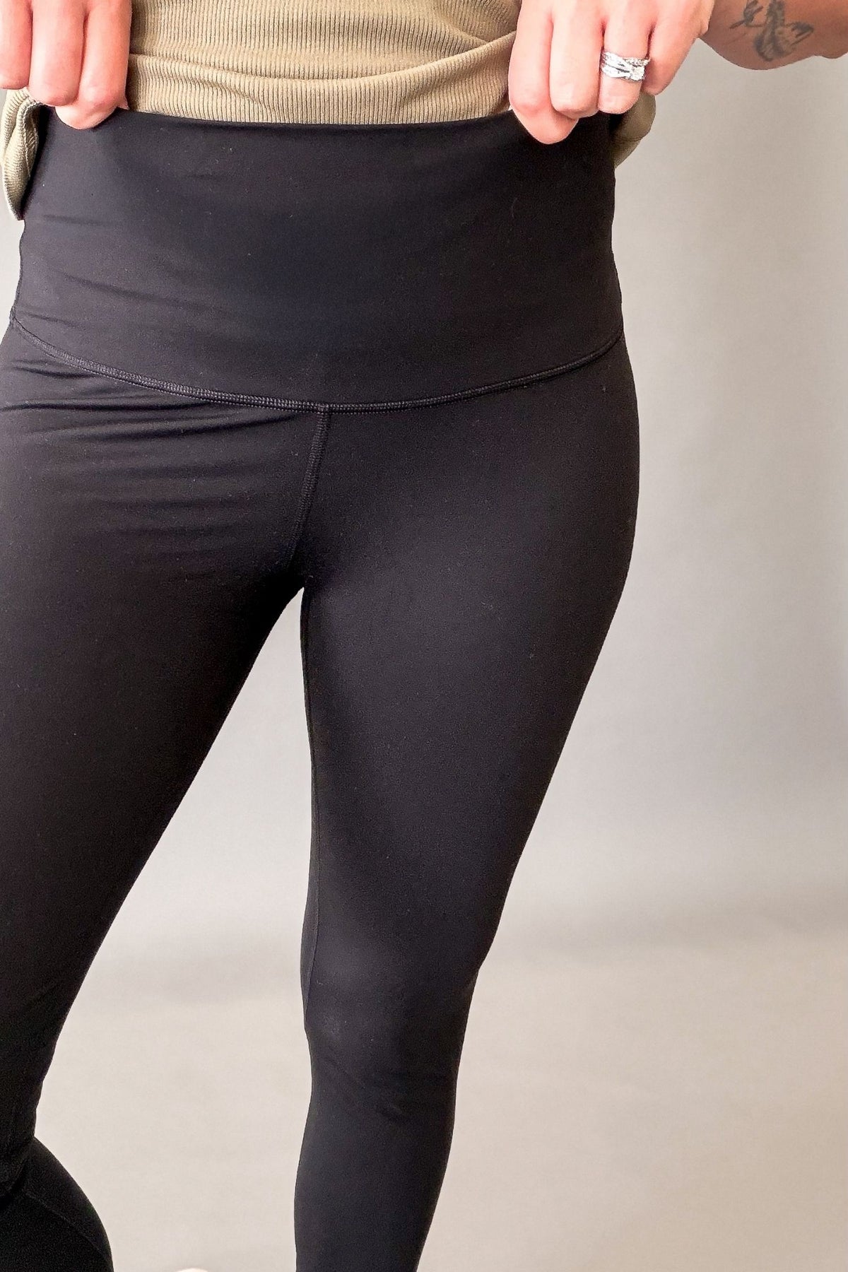 Close-up of a person wearing black high-waisted leggings, showing the waistband and upper legs against a plain light gray studio background.