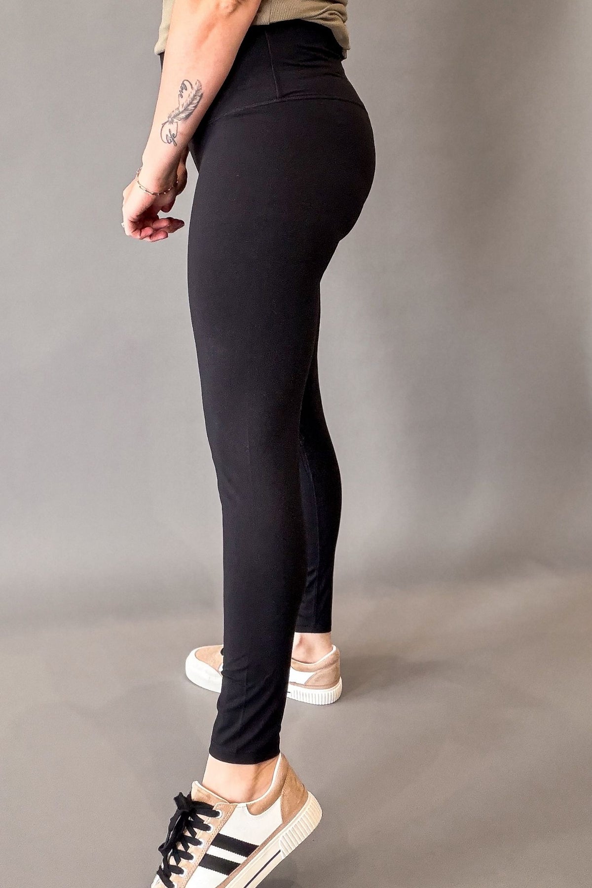 A person wearing black high-waisted leggings, shown from the side and back against a plain gray studio background. The person is also wearing a beige top and beige-and-white sneakers.