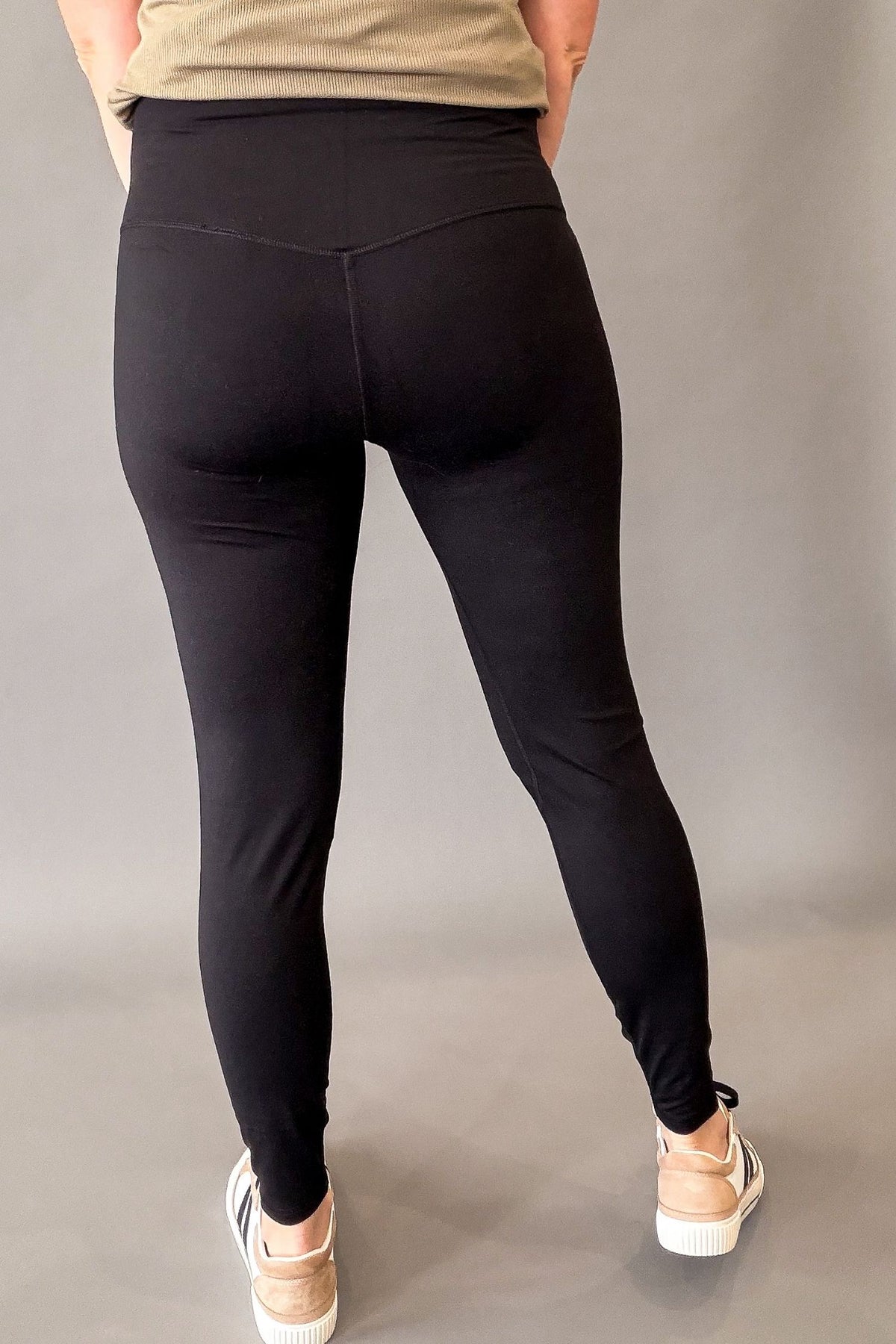 Rear view of a person wearing black high-waisted leggings, paired with a beige top and beige sneakers, against a plain gray studio background.