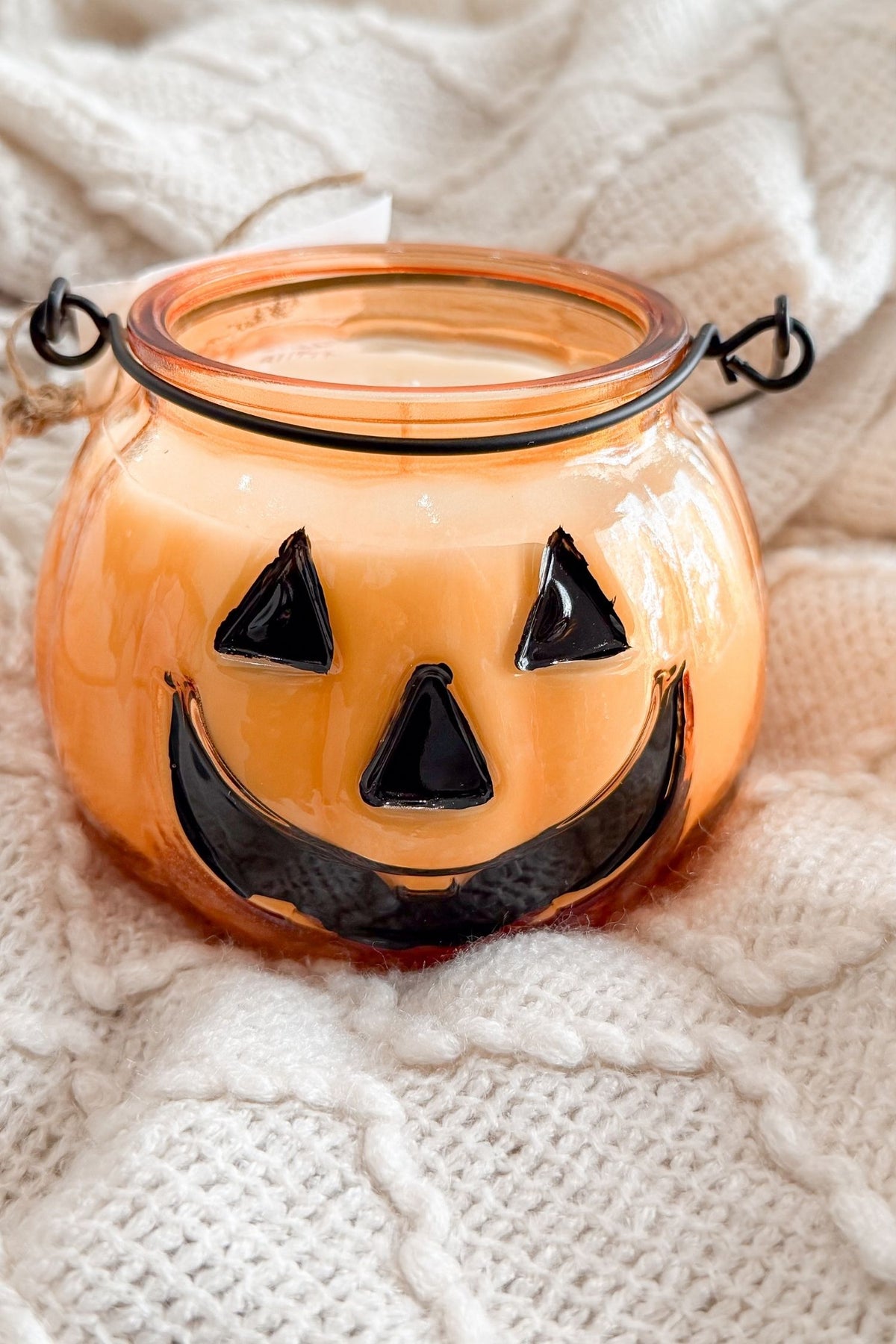 An orange pumpkin-shaped glass candle holder with a black metal handle and a black jack-o'-lantern face, resting on a white textured knit blanket.