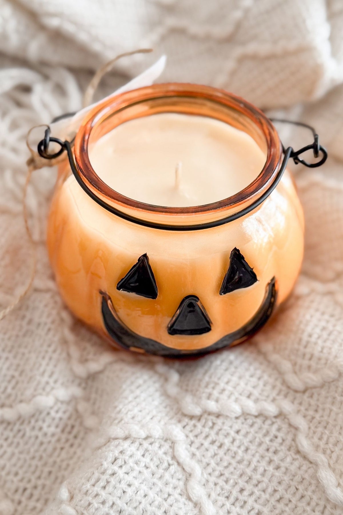 An orange pumpkin-shaped candle holder with a black hand-painted jack-o'-lantern face, filled with white wax, resting on a textured cream knit blanket.