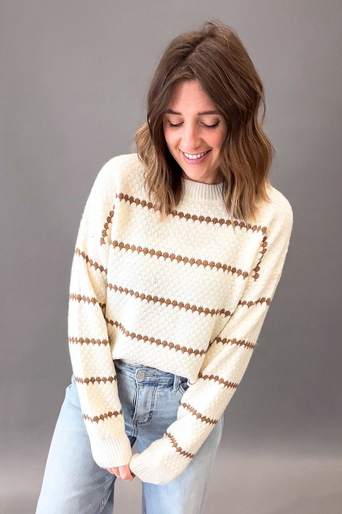 A woman wearing a cream-colored knit sweater with camel honeycomb patterns, paired with light blue jeans, posing against a solid gray studio background.
