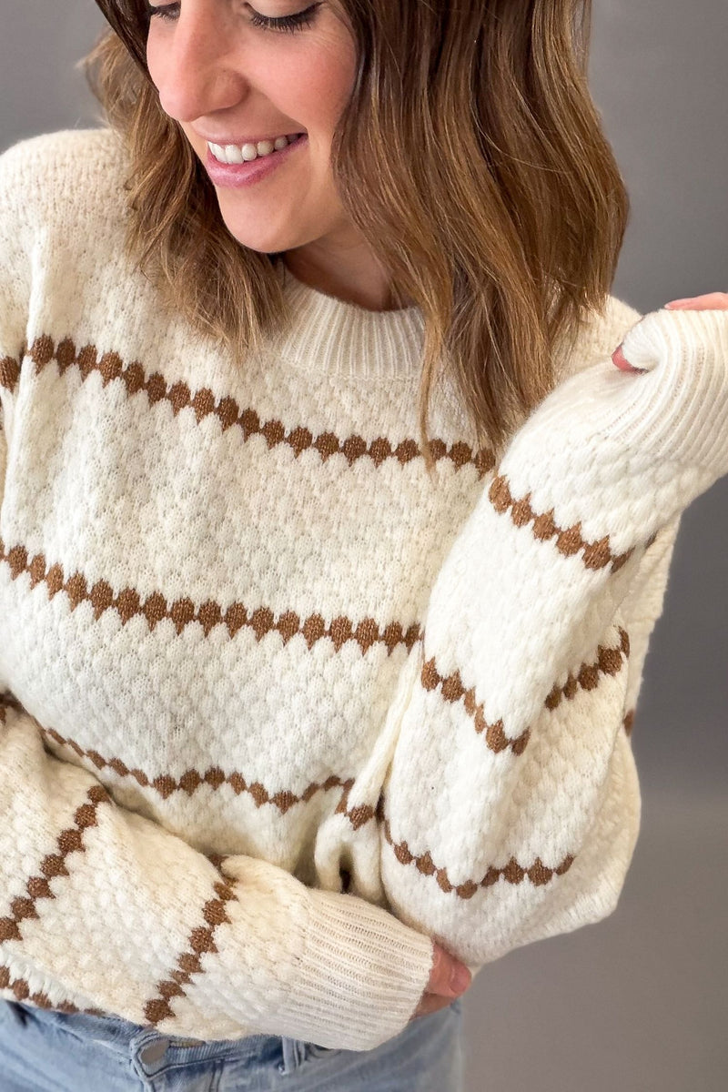 A woman wearing a camel knit sweater with brown honeycomb stripes, paired with blue jeans, shown in a close-up shot against a neutral gray background.