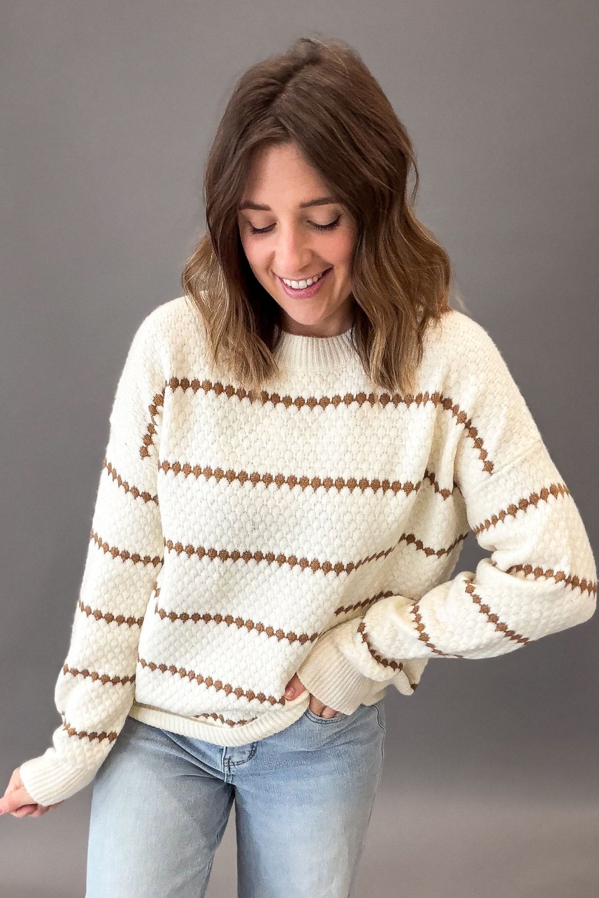 A woman wearing a camel-colored knit sweater with brown horizontal honeycomb stripes, paired with light blue jeans, standing against a solid gray studio background.