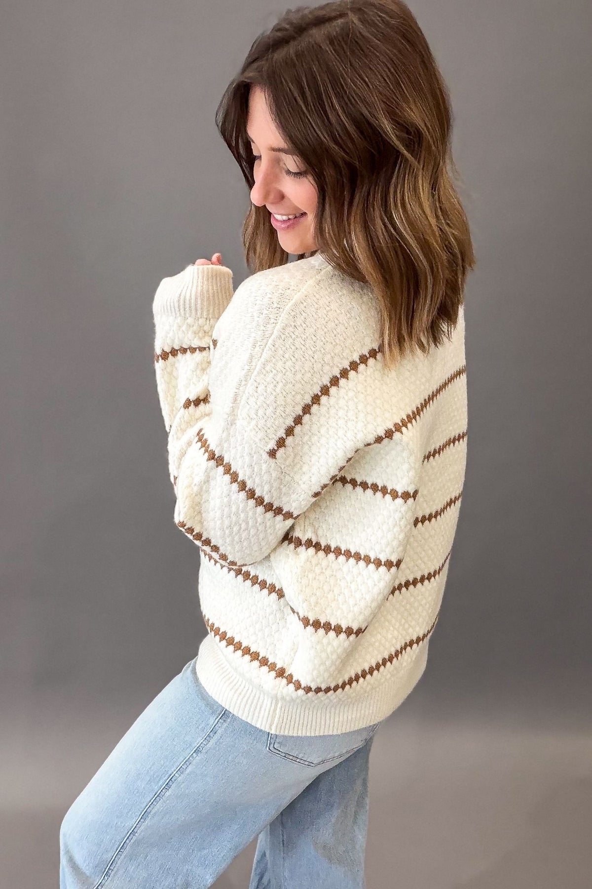 A woman wearing a Ivory-colored knit sweater with camel horizontal honeycomb stripes, paired with light blue jeans, photographed against a solid gray studio background.