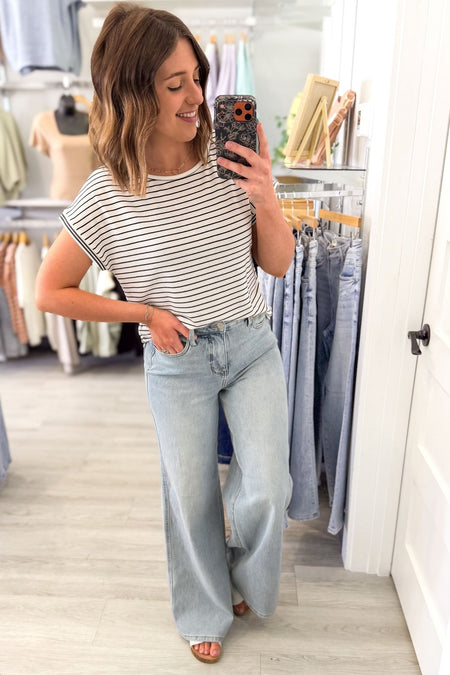 Woman taking a mirror selfie wearing a black striped shirt and light blue jeans in a clothing store.