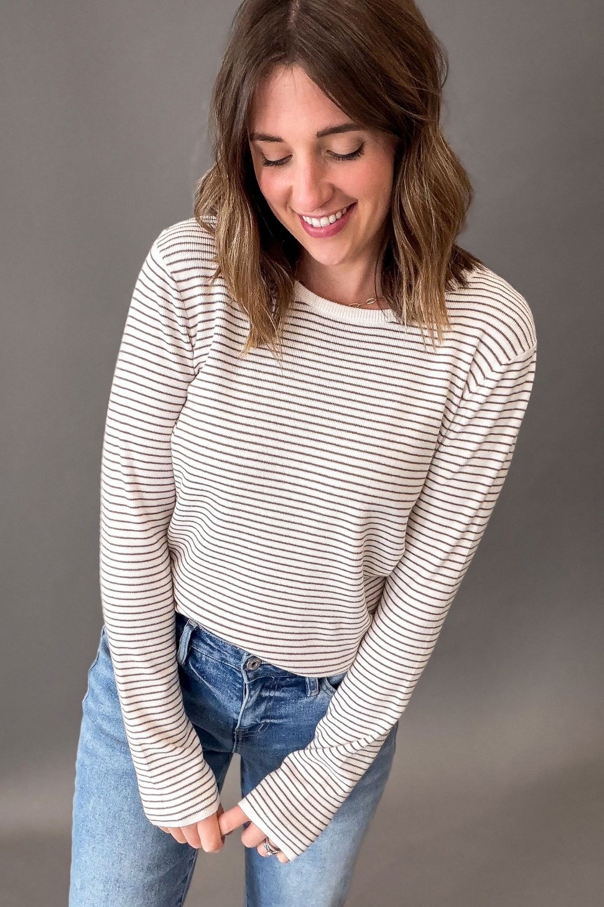 A woman wearing a ivory + mocha horizontally striped long-sleeve sweater, paired with blue jeans, posing against a solid gray studio background.