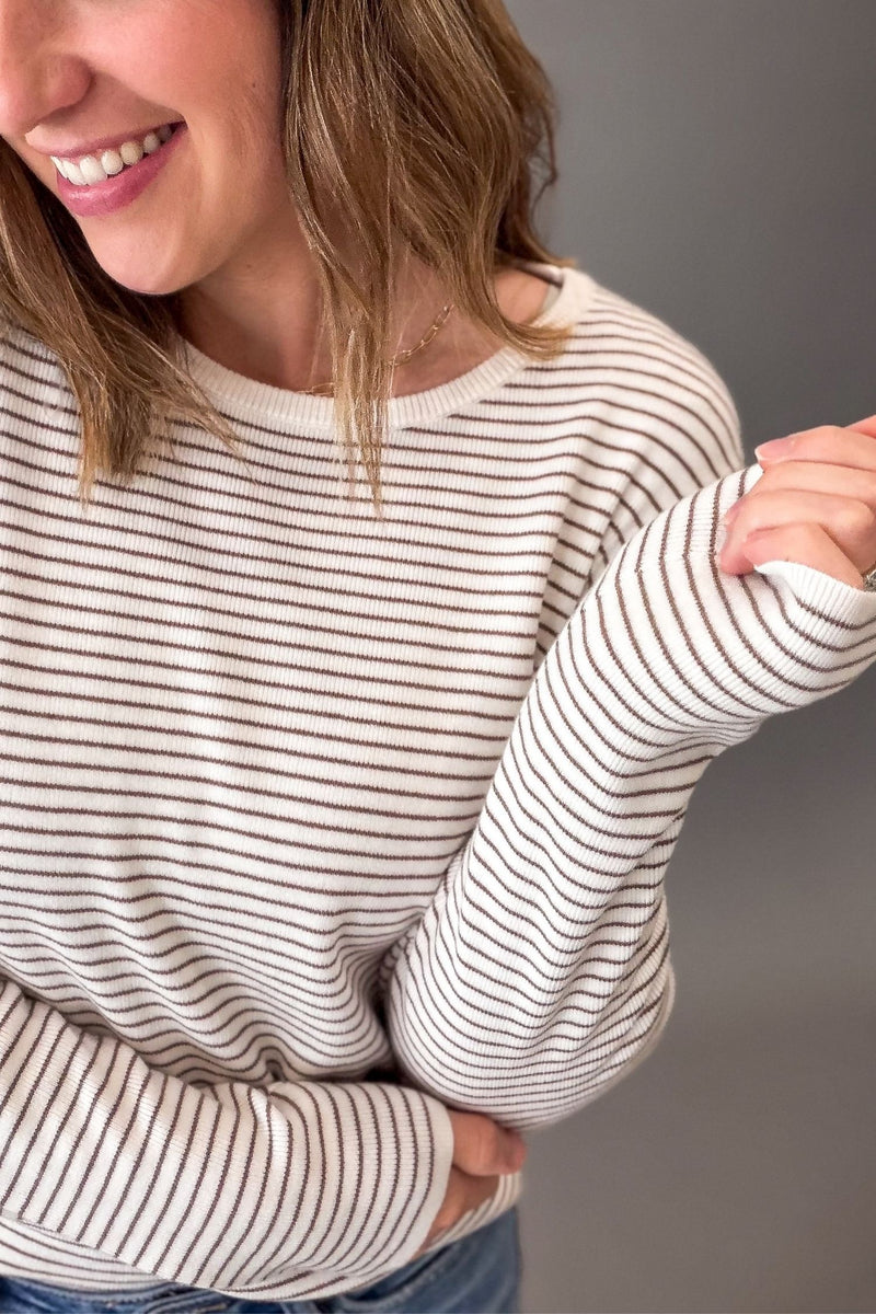A woman wearing a ivory-colored long-sleeve knit sweater with thin mocha horizontal stripes, shown from the chest up against a solid gray background.